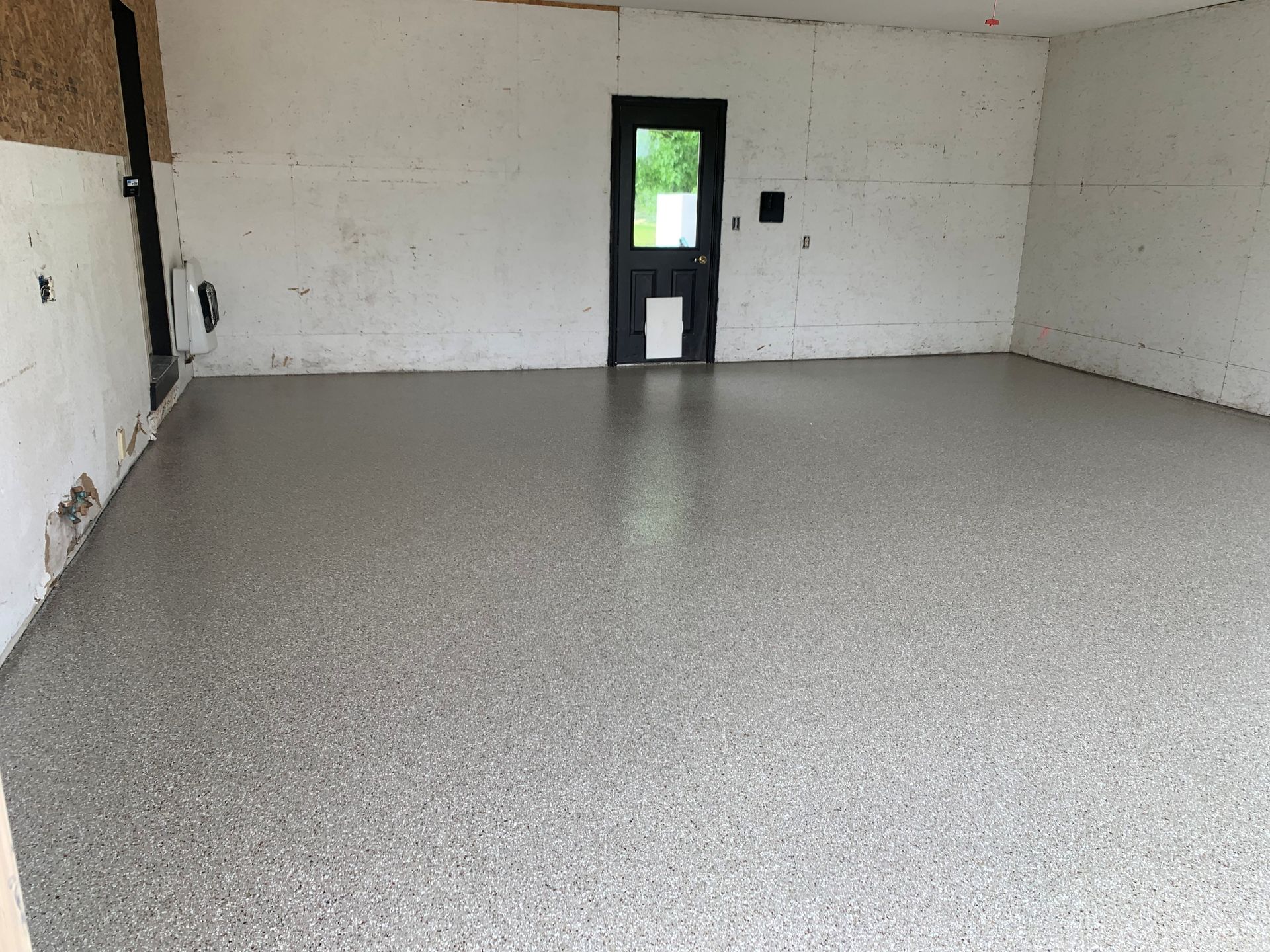Empty white room with a gray speckled floor and a small black door opening to greenery outside