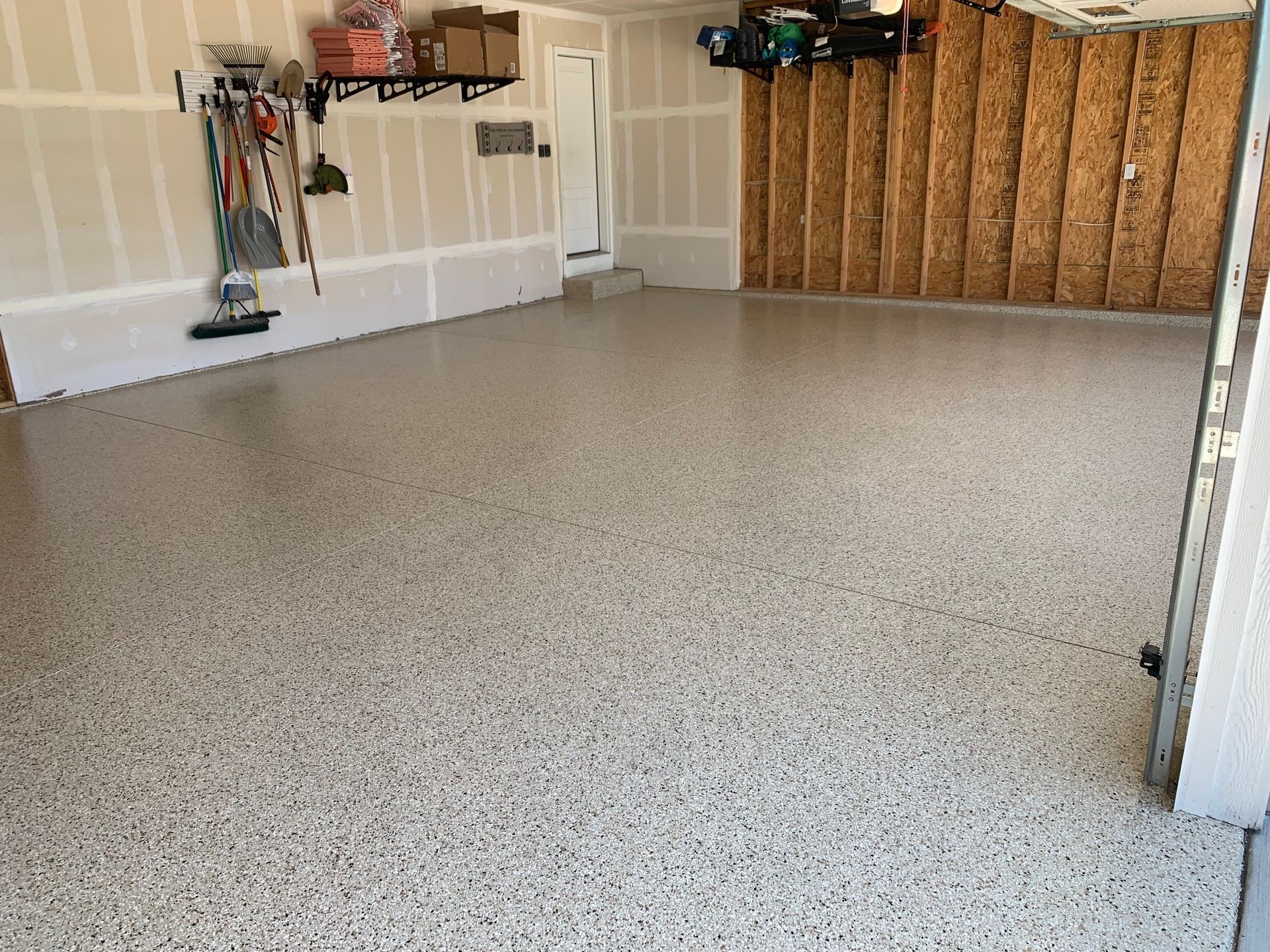 Empty garage with speckled epoxy floor, unfinished walls, and storage shelves along the back.