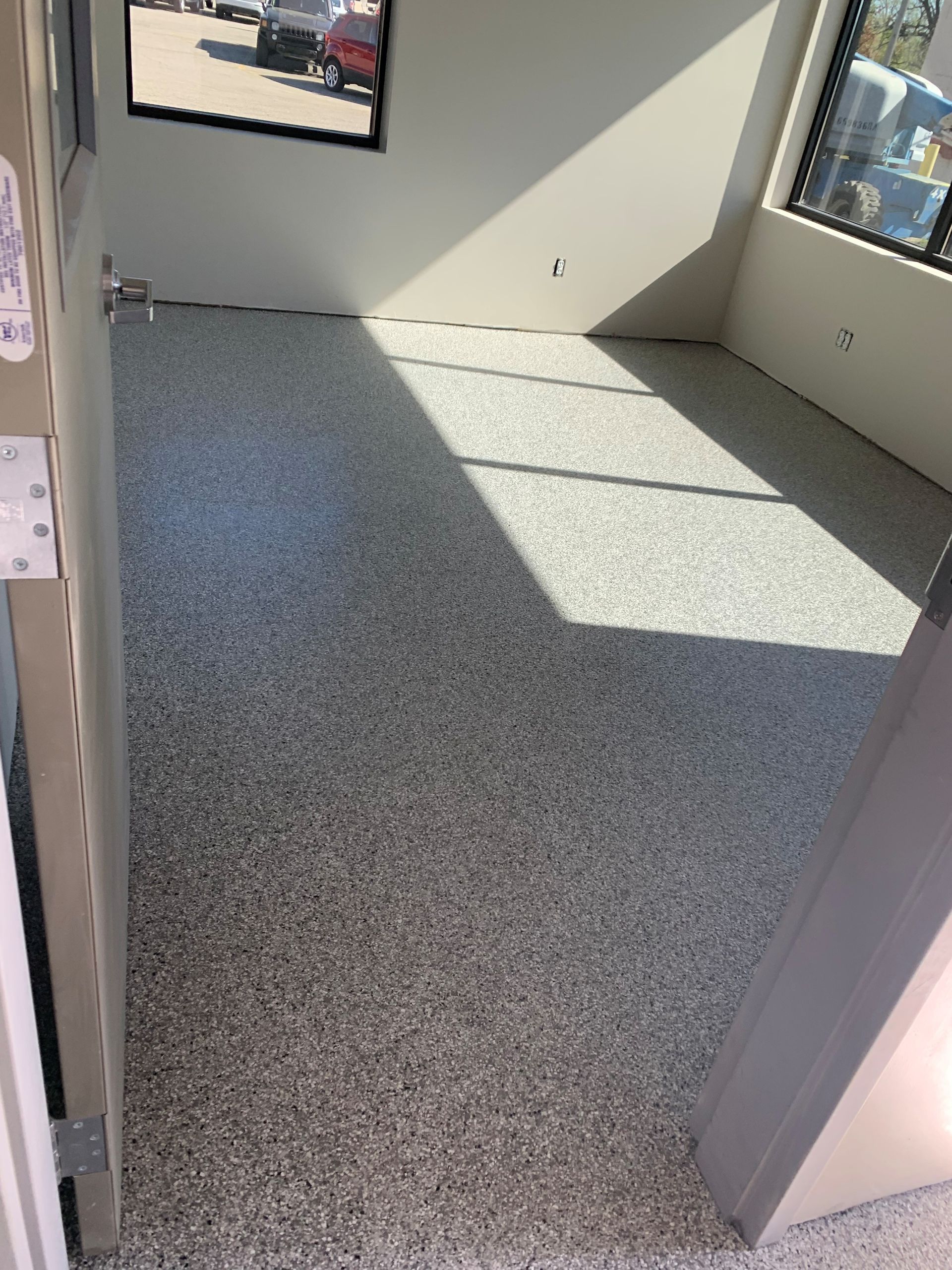 Empty sunlit room with speckled gray floor and large windows.