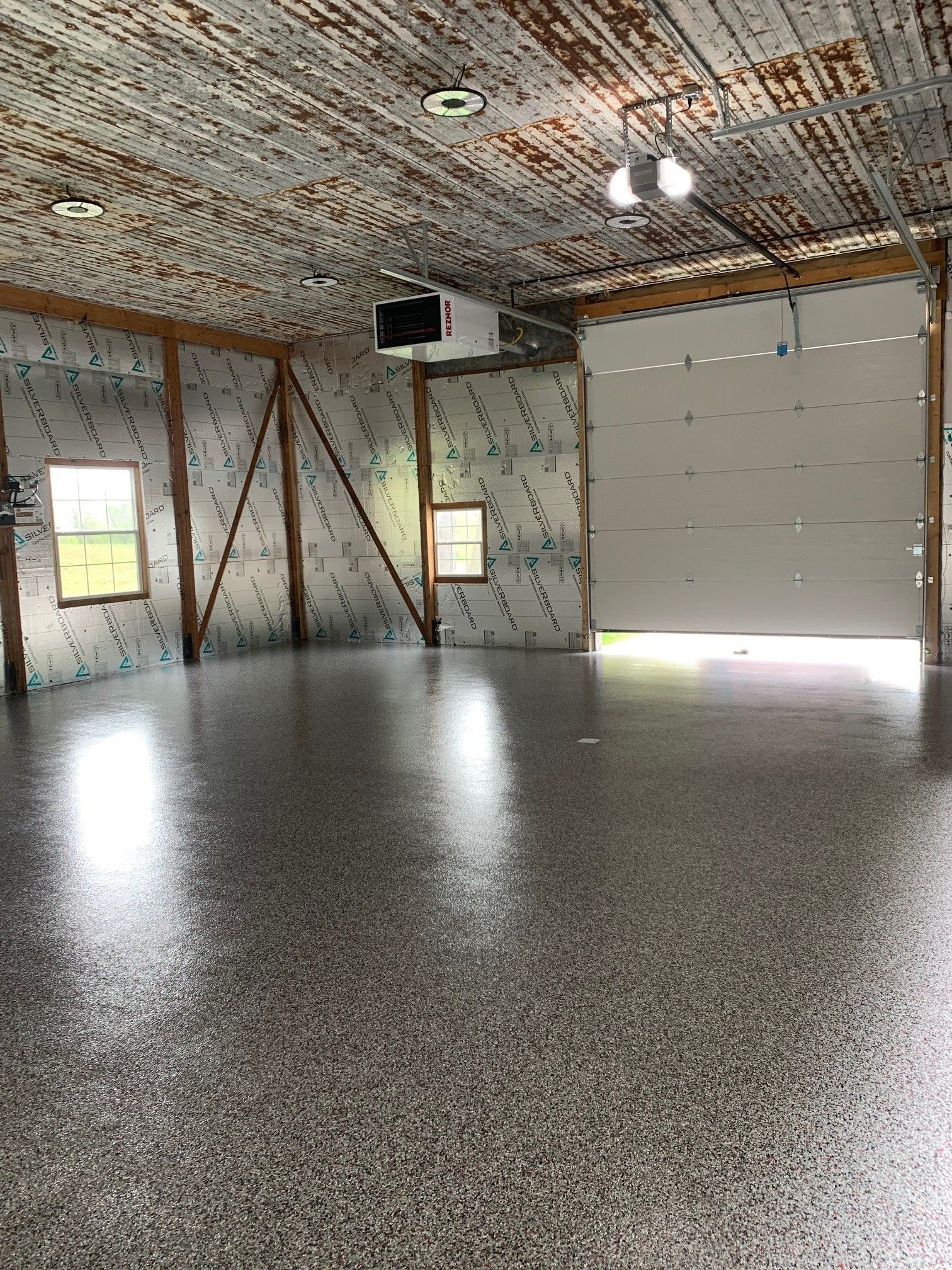Empty garage with speckled concrete floor, metal walls, windows, and a large overhead door open to daylight