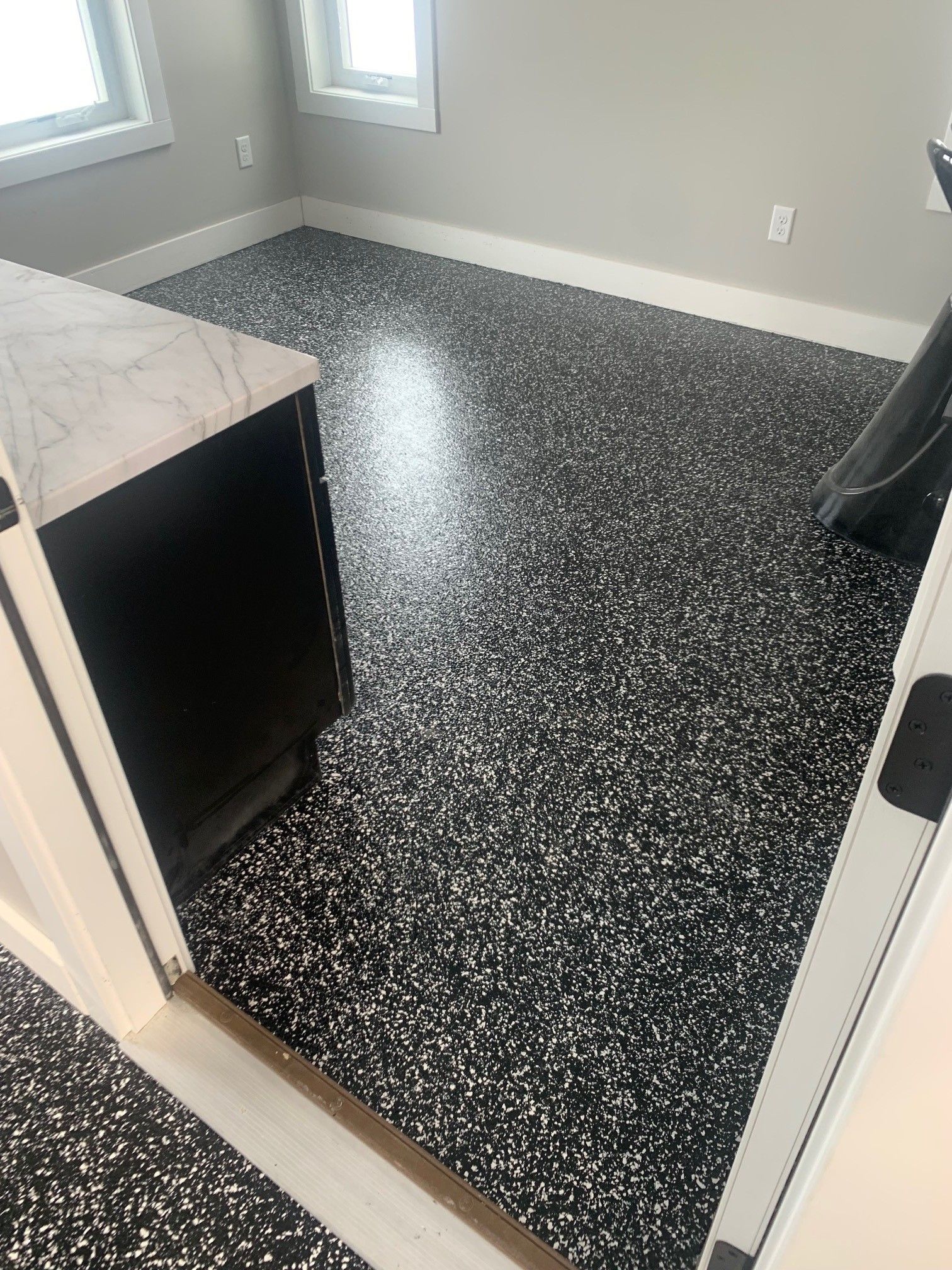 Small empty room with black speckled floor, light gray walls, and two windows.
