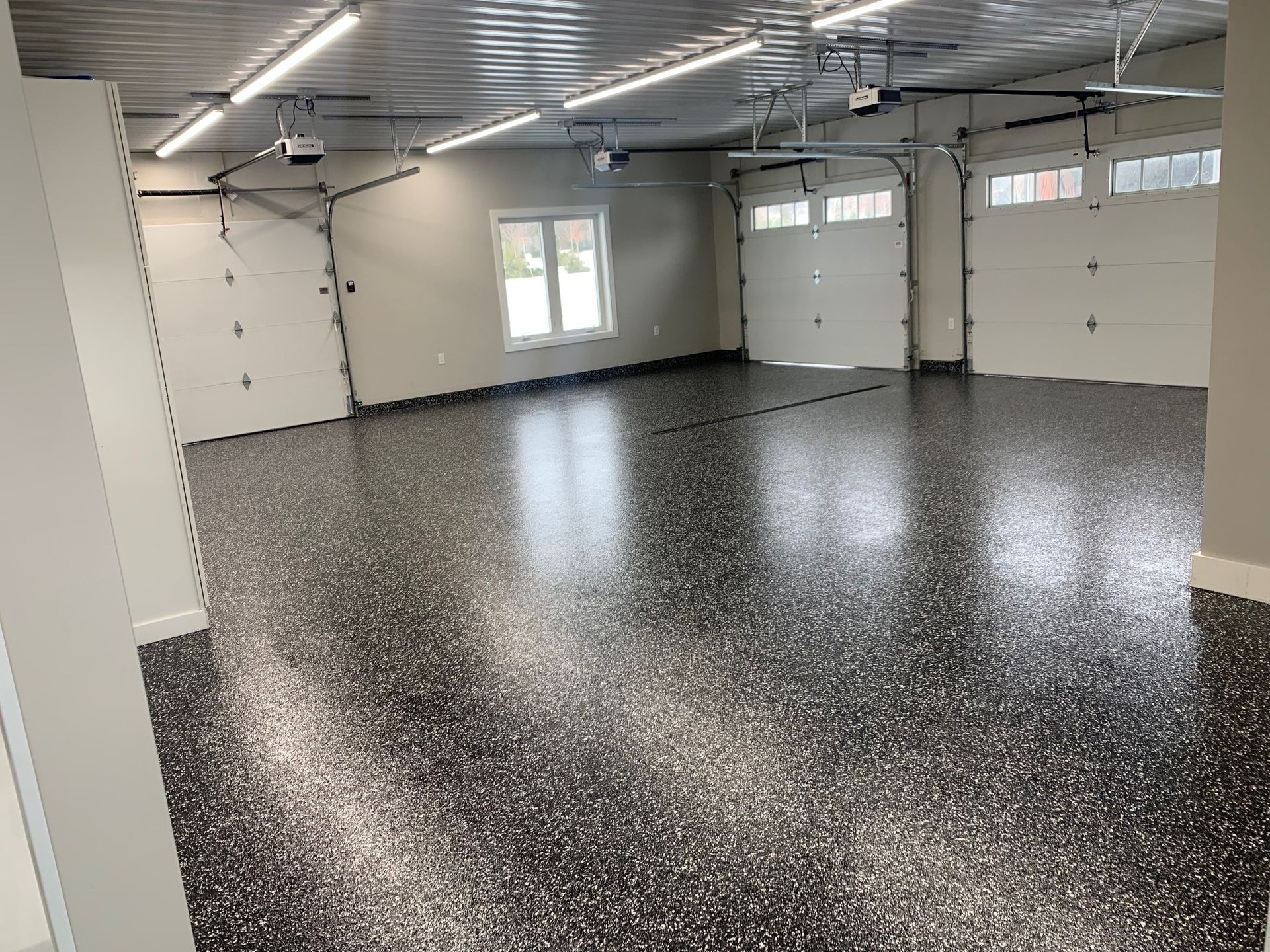 Empty garage with speckled epoxy floor, white walls, garage doors, and ceiling lights.