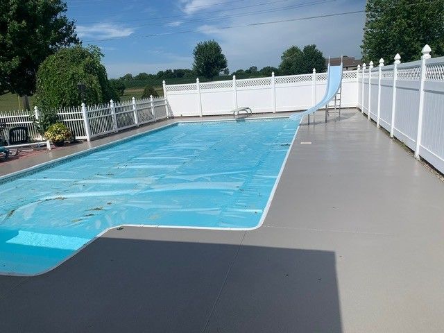 Covered backyard swimming pool beside a concrete deck and white fence under a blue sky