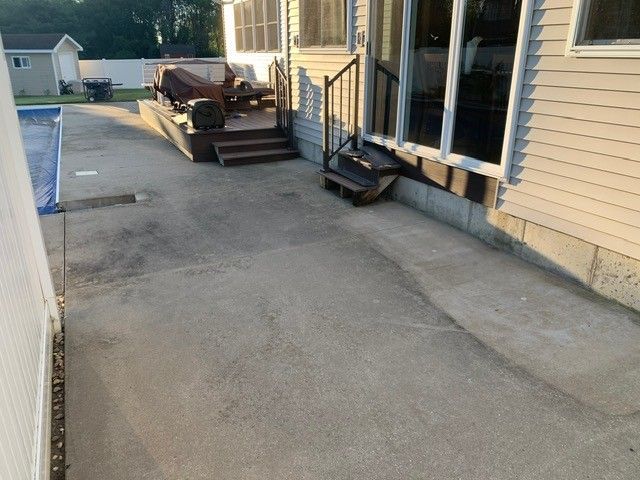 Concrete side yard beside a beige house, with stairs, outdoor chairs, and a driveway in the background
