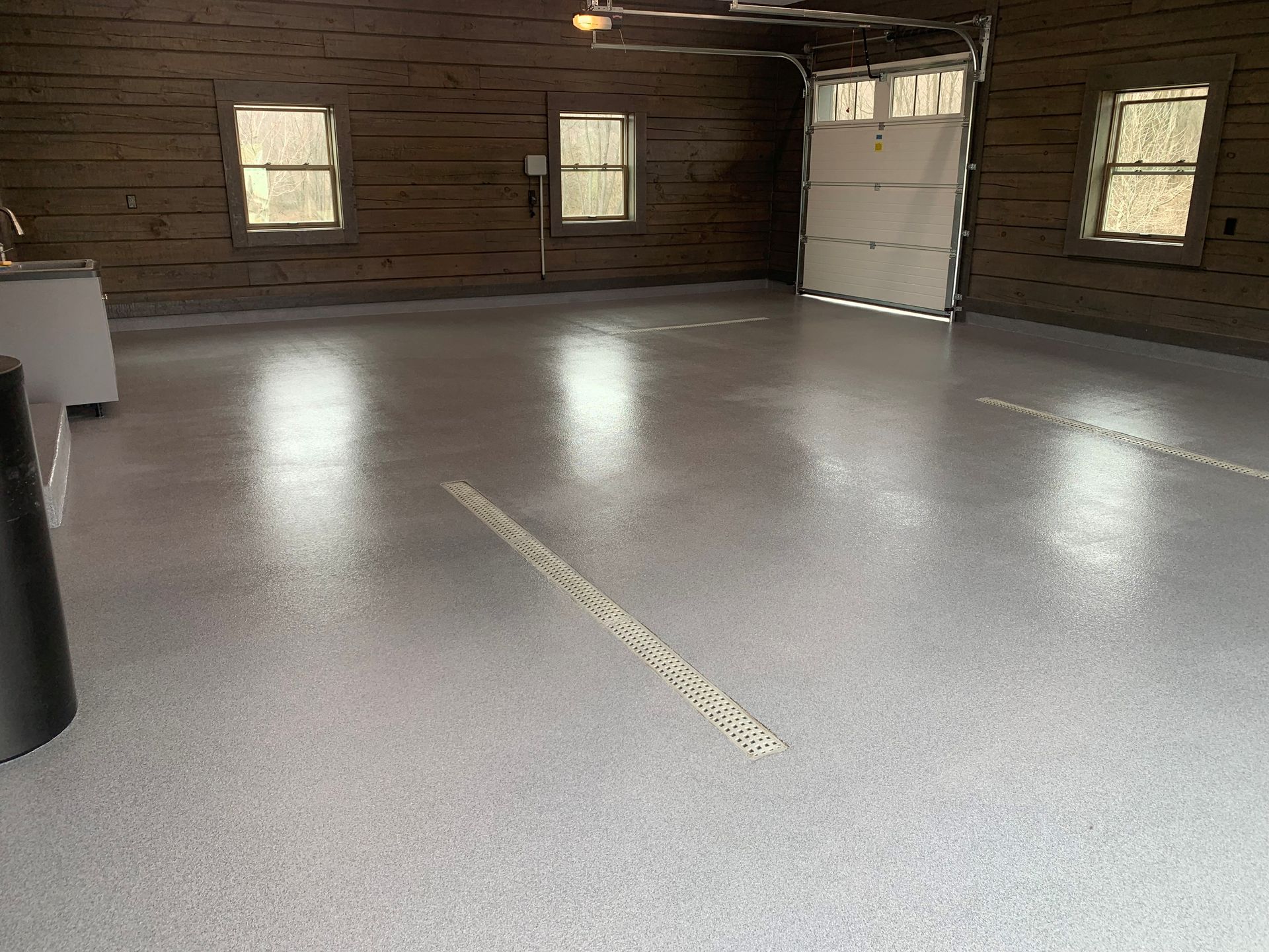 Empty garage with gray epoxy floor, brown wood walls, small windows, and a white overhead door.