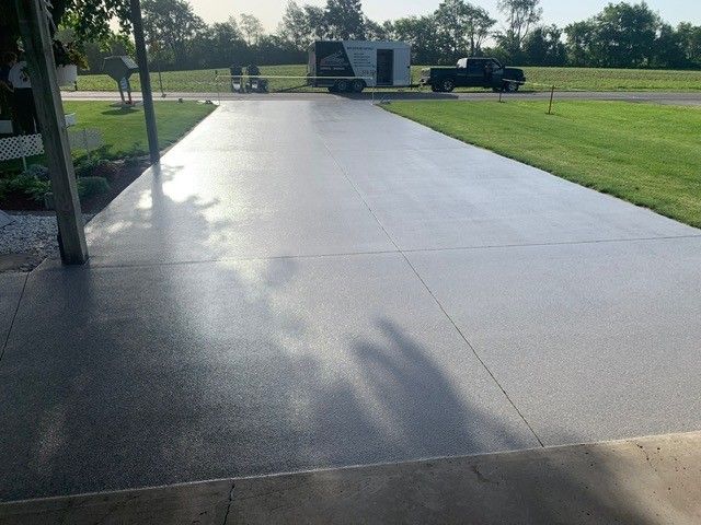 Wide concrete walkway in a park, with green grass and trees under bright sunlight.