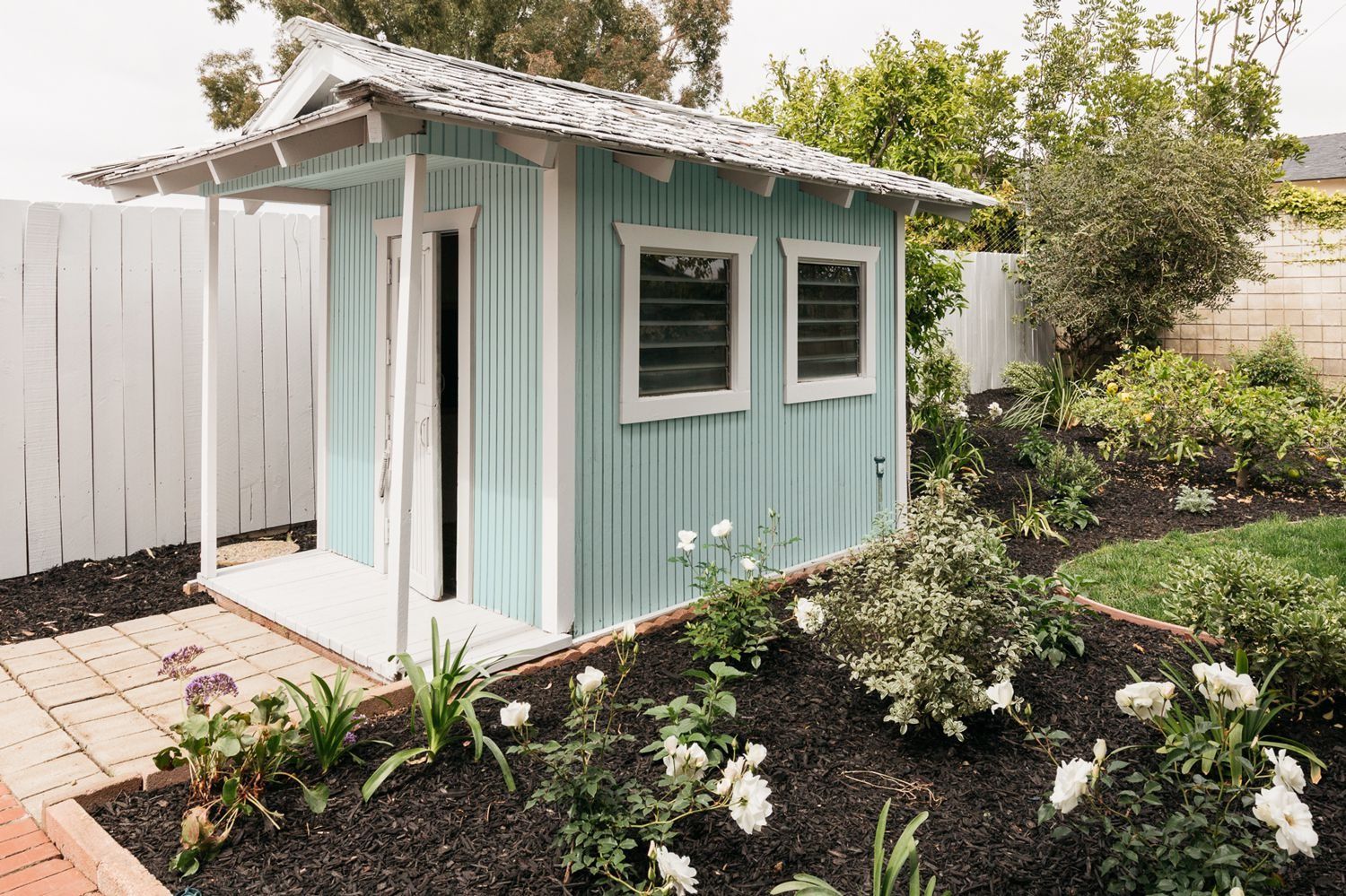 A small, light-blue backyard shed with a porch and white trim, set in a garden with dark mulch and white flowers.