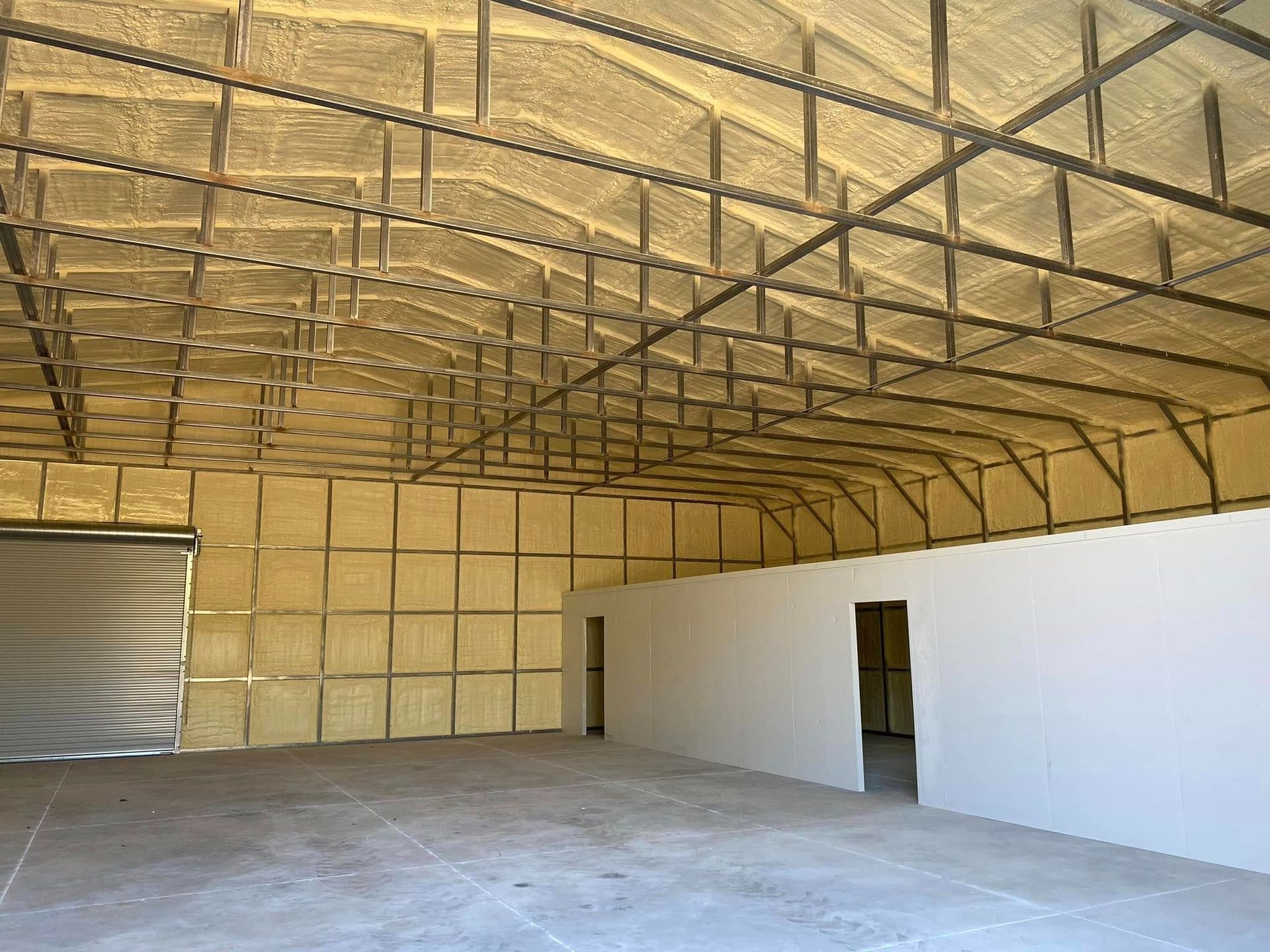 Large industrial warehouse interior with exposed steel roof trusses insulated with yellow spray foam and white walls.