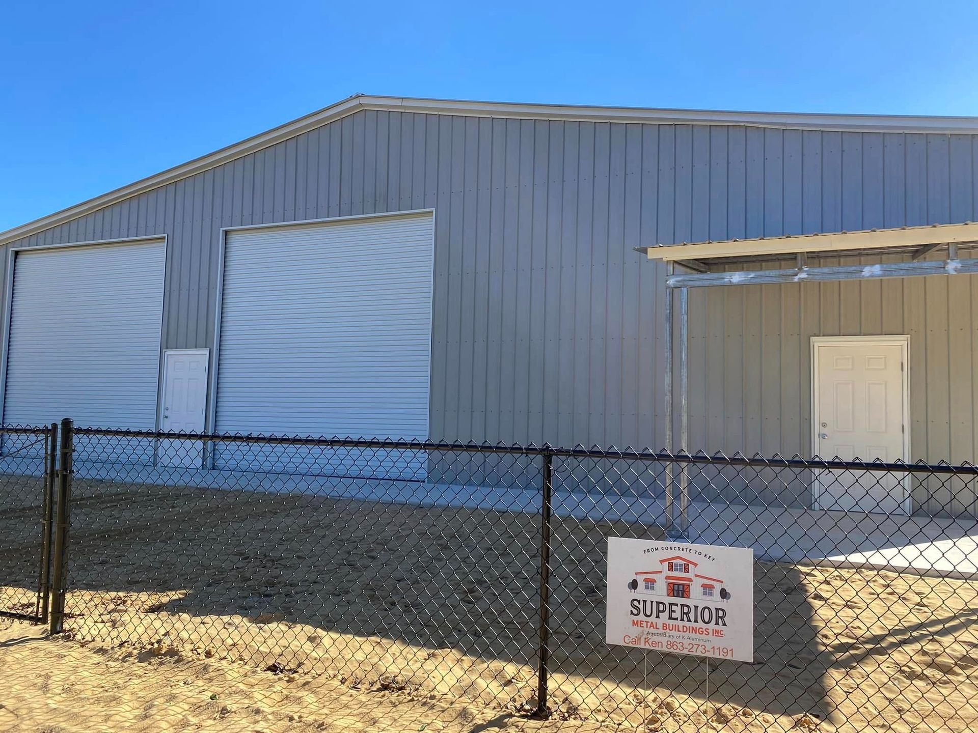 A grey metal warehouse building with two roll-up doors and a white pedestrian door, fronted by a black chain-link fence.