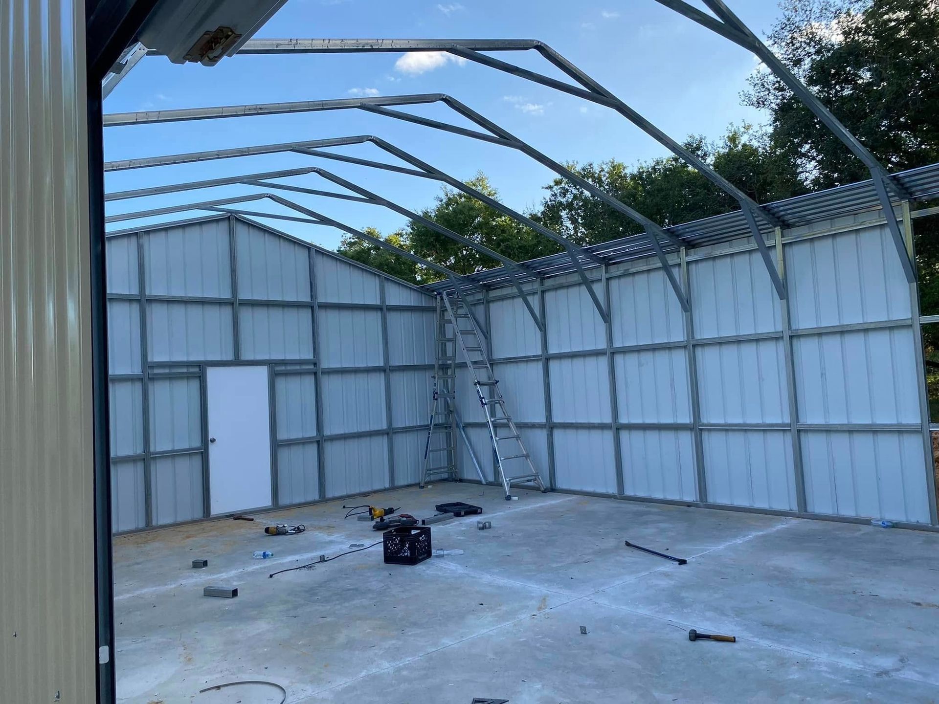 A partially constructed metal building with wall panels installed, an open roof frame, and tools scattered on a concrete floor.