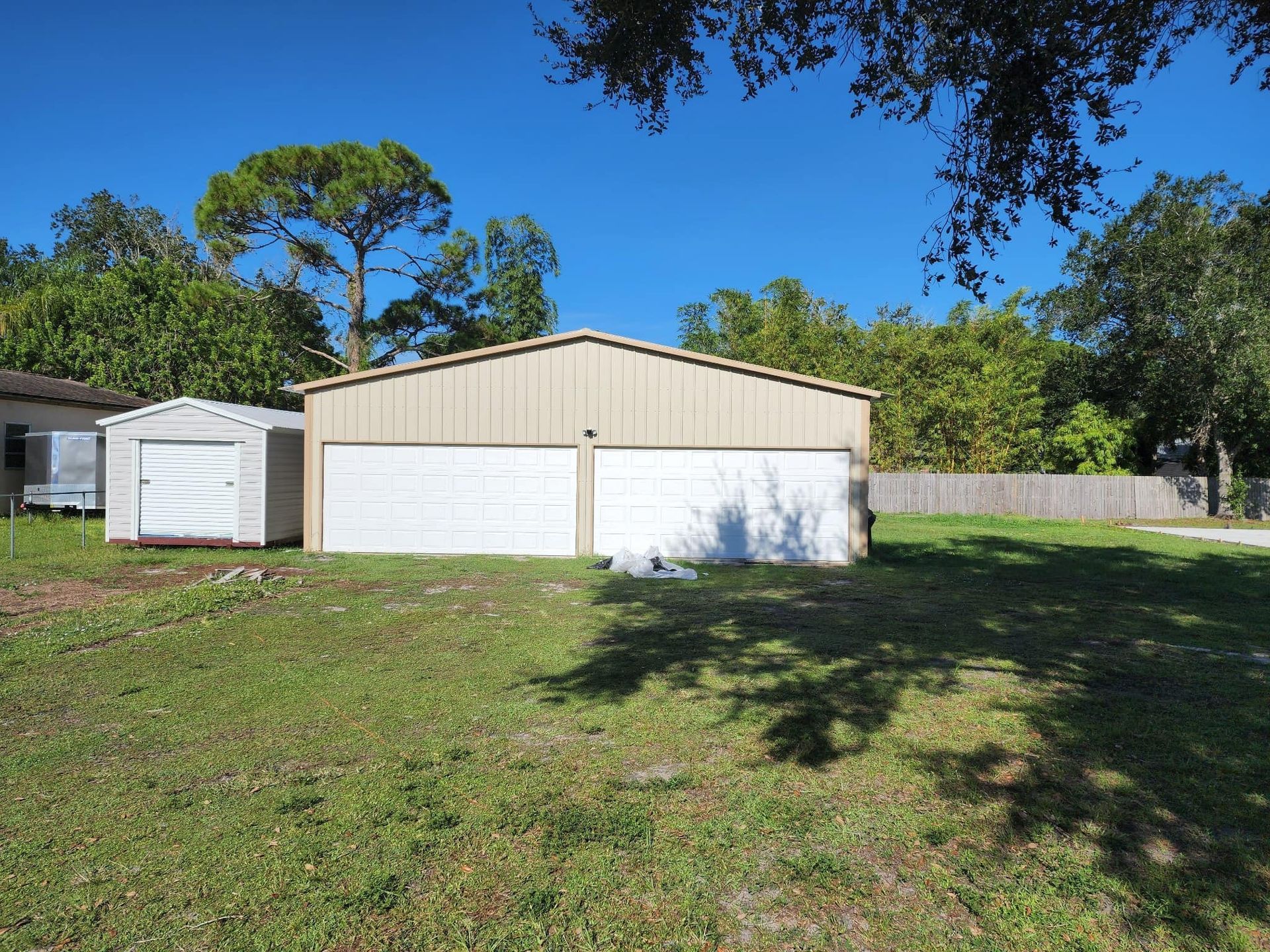 A two-car tan garage with white doors and a small shed in a grassy yard under a clear blue sky.