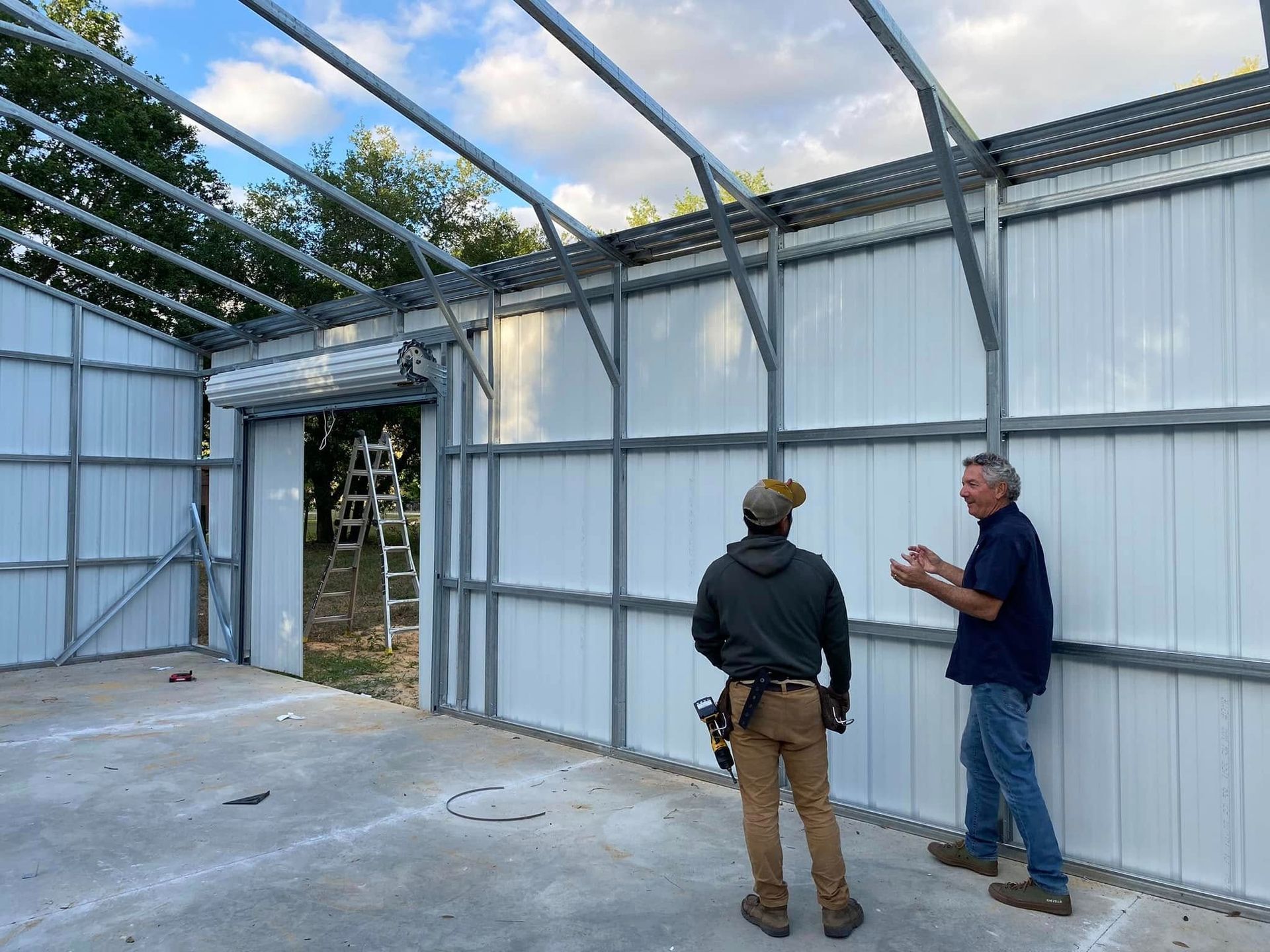 Two people stand inside a large metal-framed building under construction, discussing the installation of wall panels.