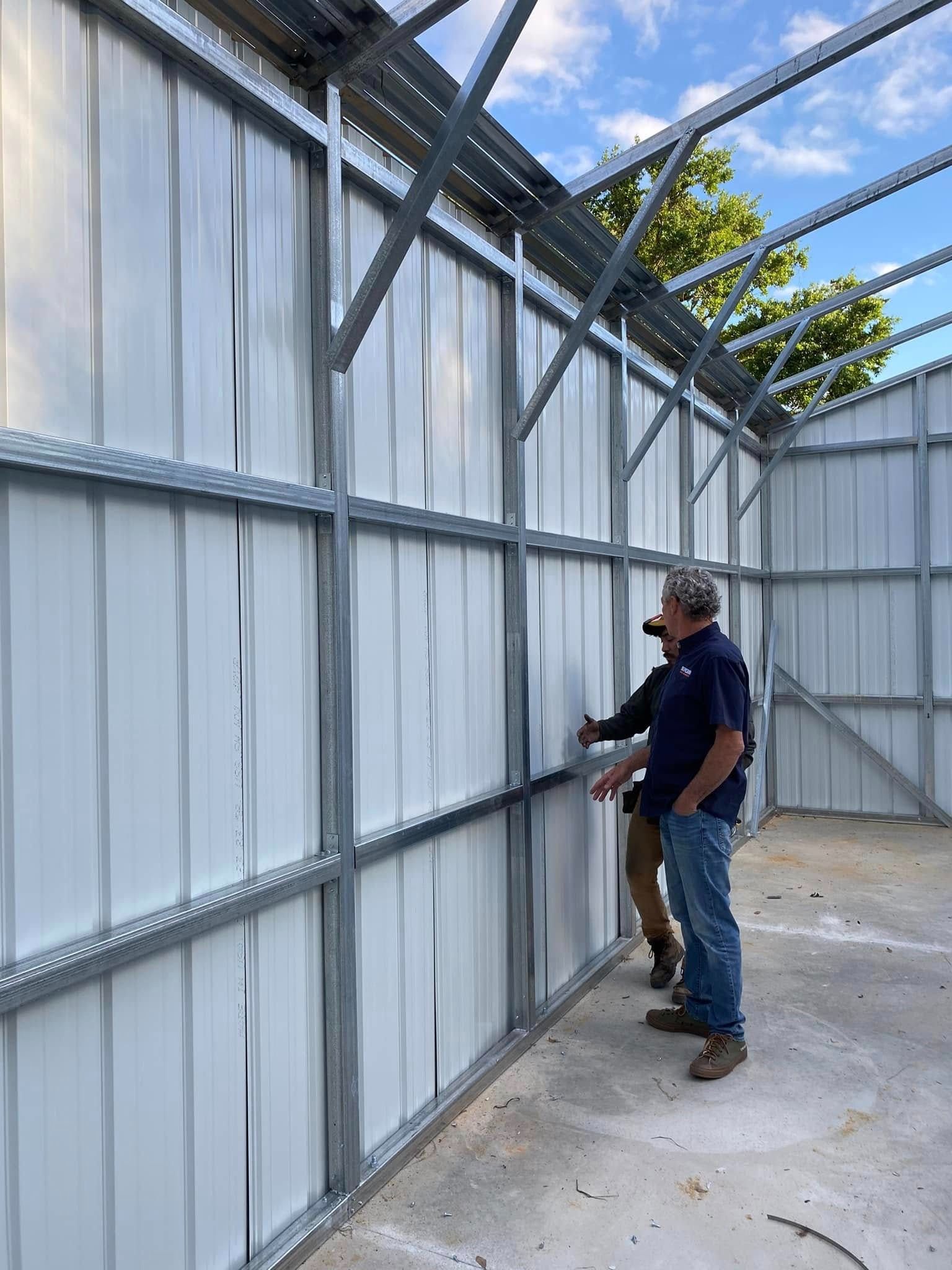 Two individuals inspect the metal wall panels and framing structure of an unfinished metal building.