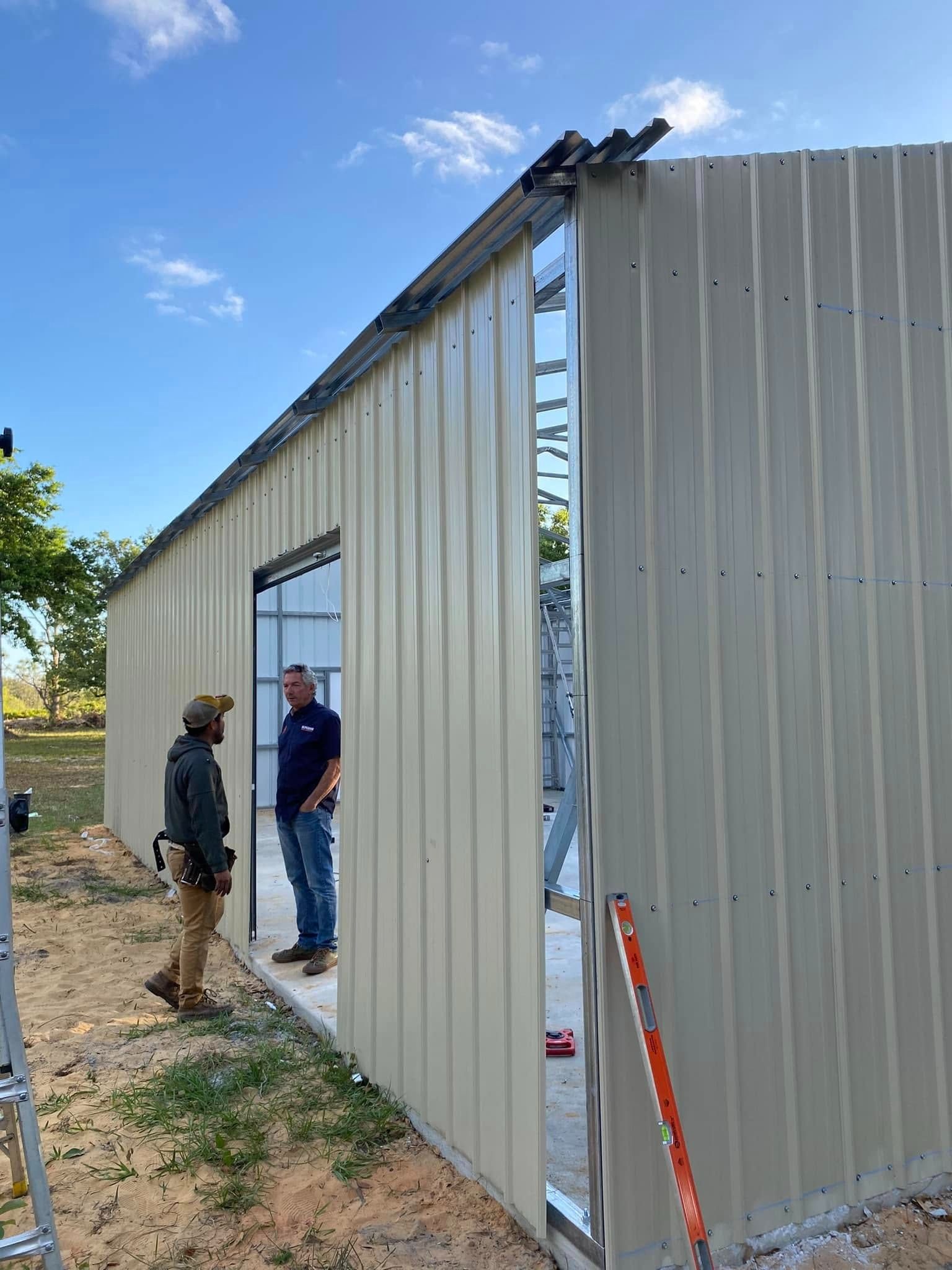 Two workers stand near the doorway of a partially constructed metal building on a sunny day.