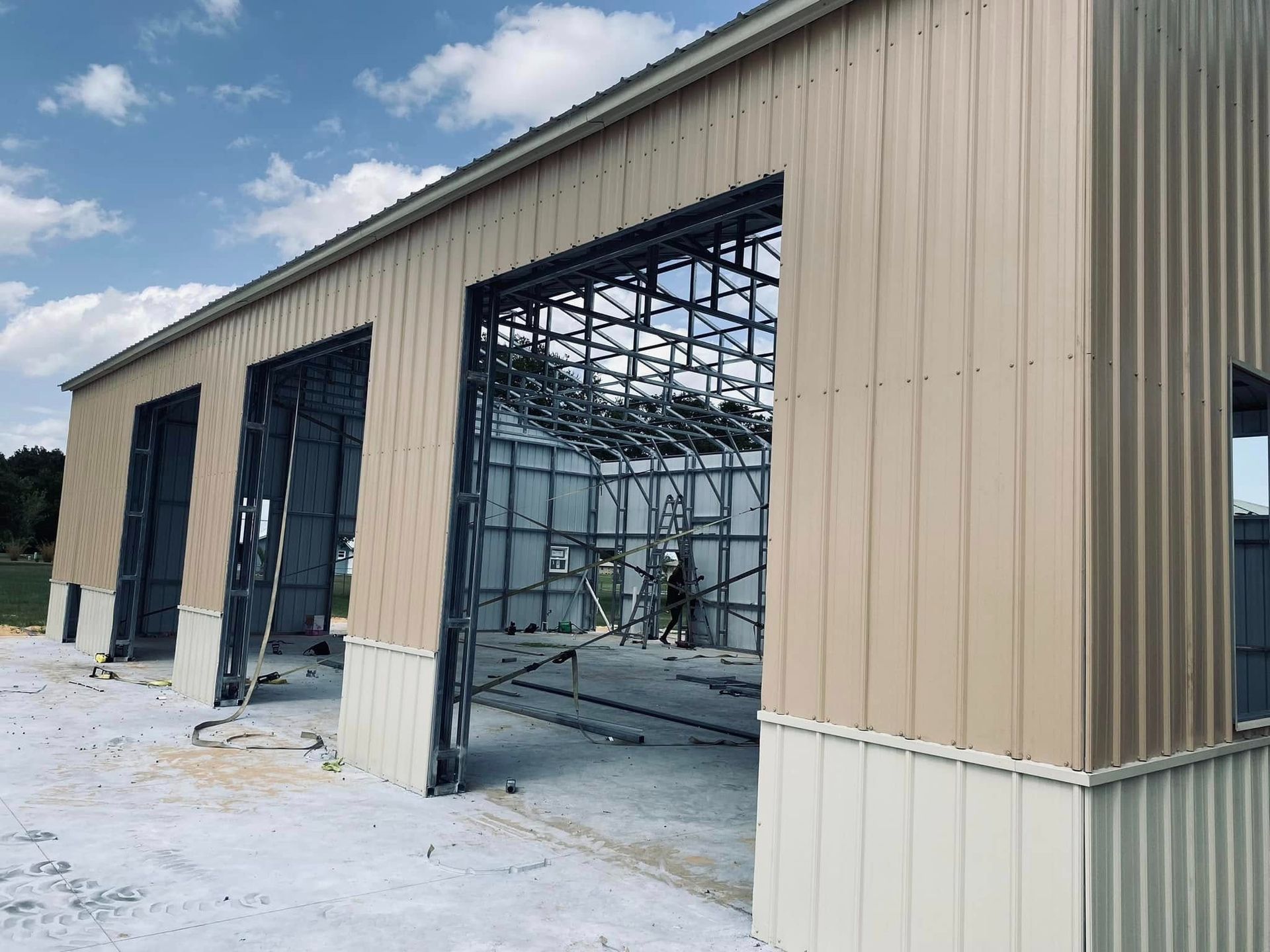 A partially constructed metal building with tan siding, large open garage door frames, and an exposed steel roof structure.
