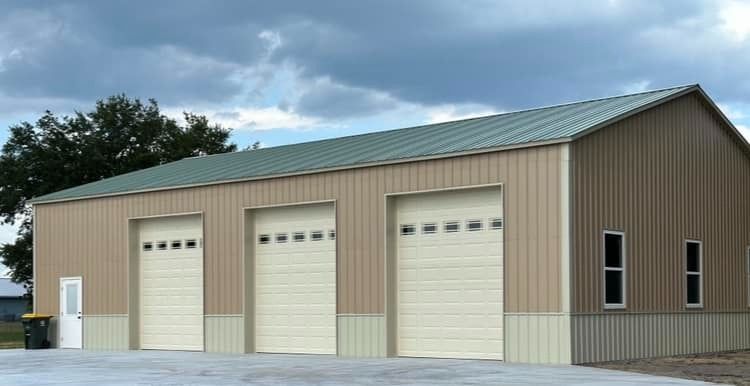 A tan metal pole barn with a green roof, three garage doors with windows, and a white entry door on a concrete pad.