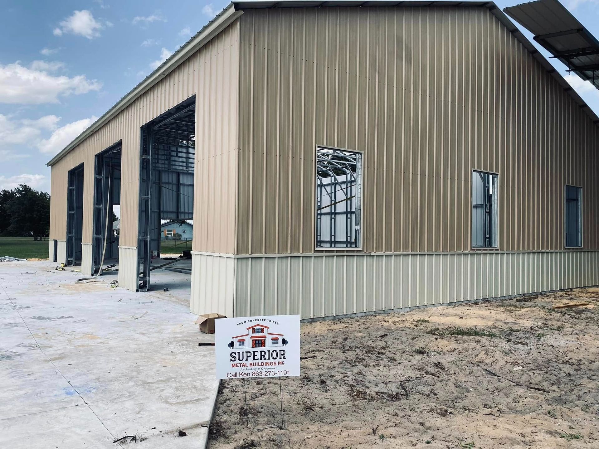 A partially constructed tan metal building with open garage bays, exterior windows, and a small yard sign in the foreground.