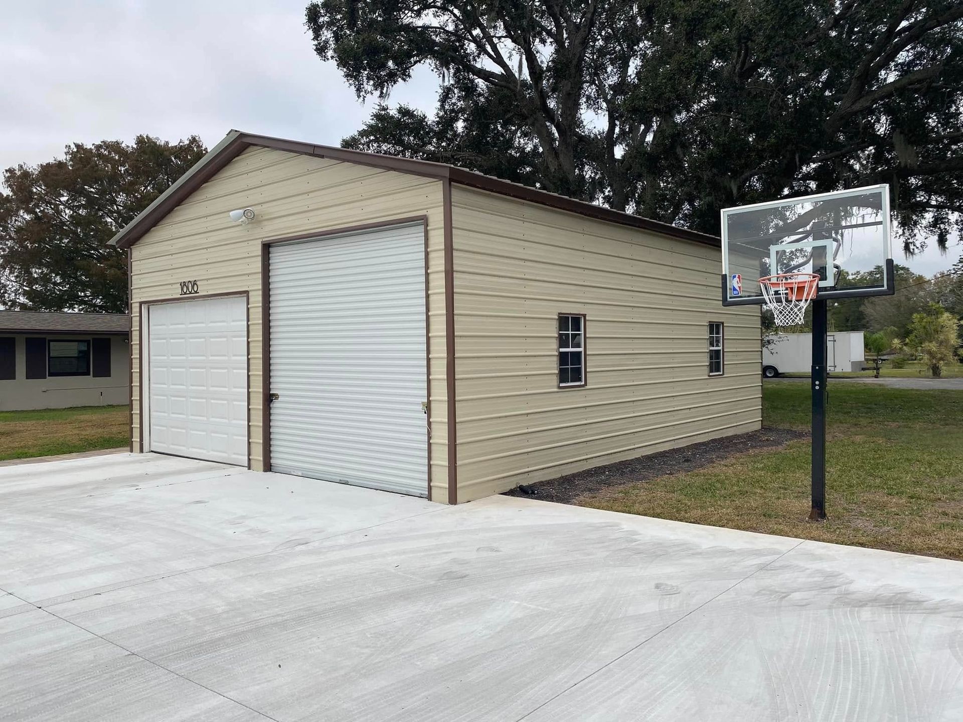 A tan metal garage with two roll-up doors and two windows, located next to a basketball hoop on a concrete driveway.