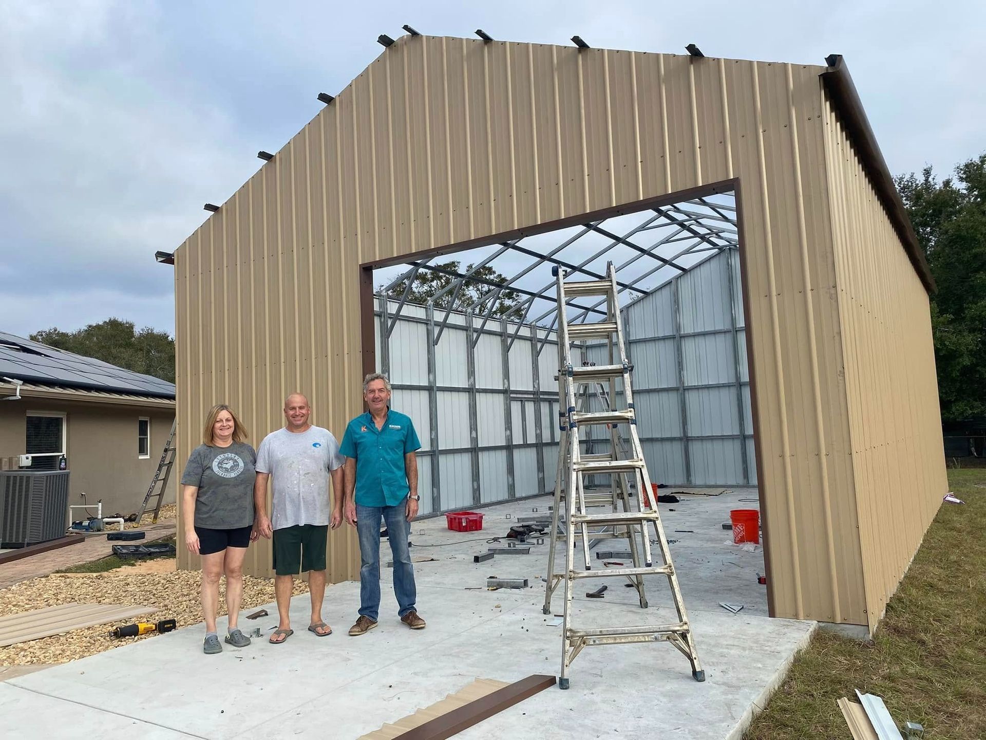Three people stand outside a tan metal garage building under construction, with a ladder positioned in the doorway.