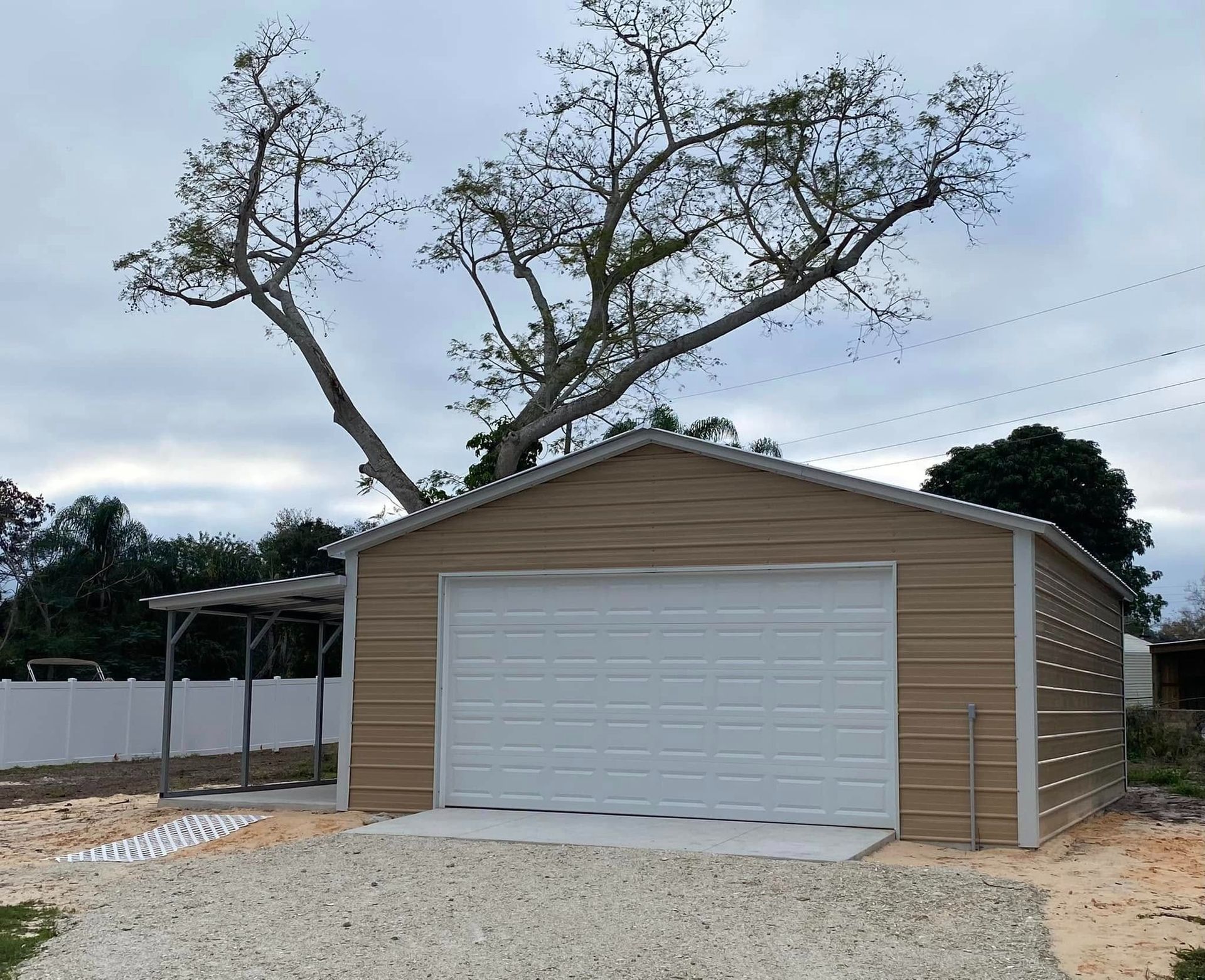 A tan metal garage with a white roll-up door and an attached side carport sits on a gravel lot under a large leafless tree.