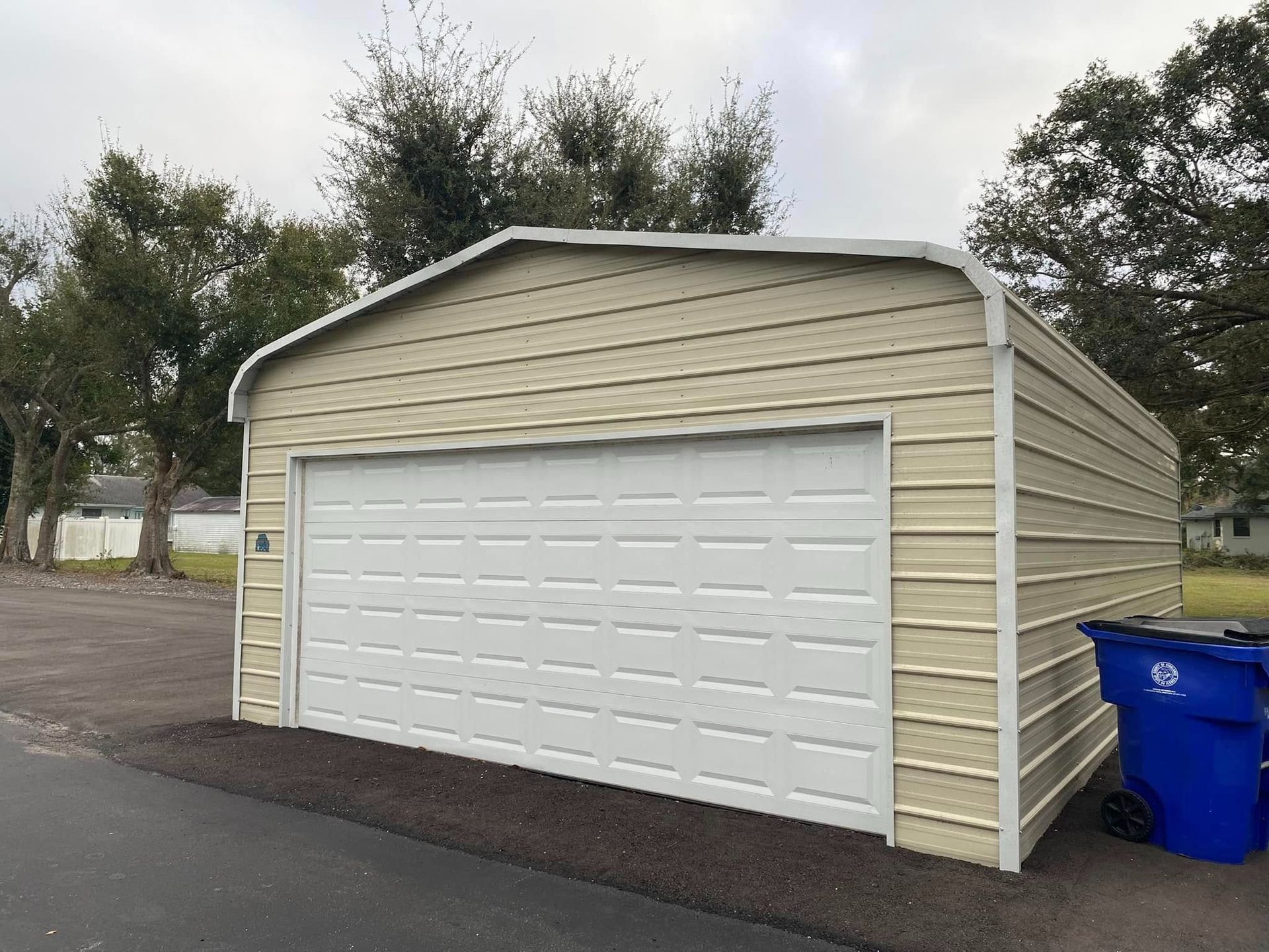 A detached, beige metal garage with a white roll-up door and rounded roof, situated outdoors next to a blue trash bin.