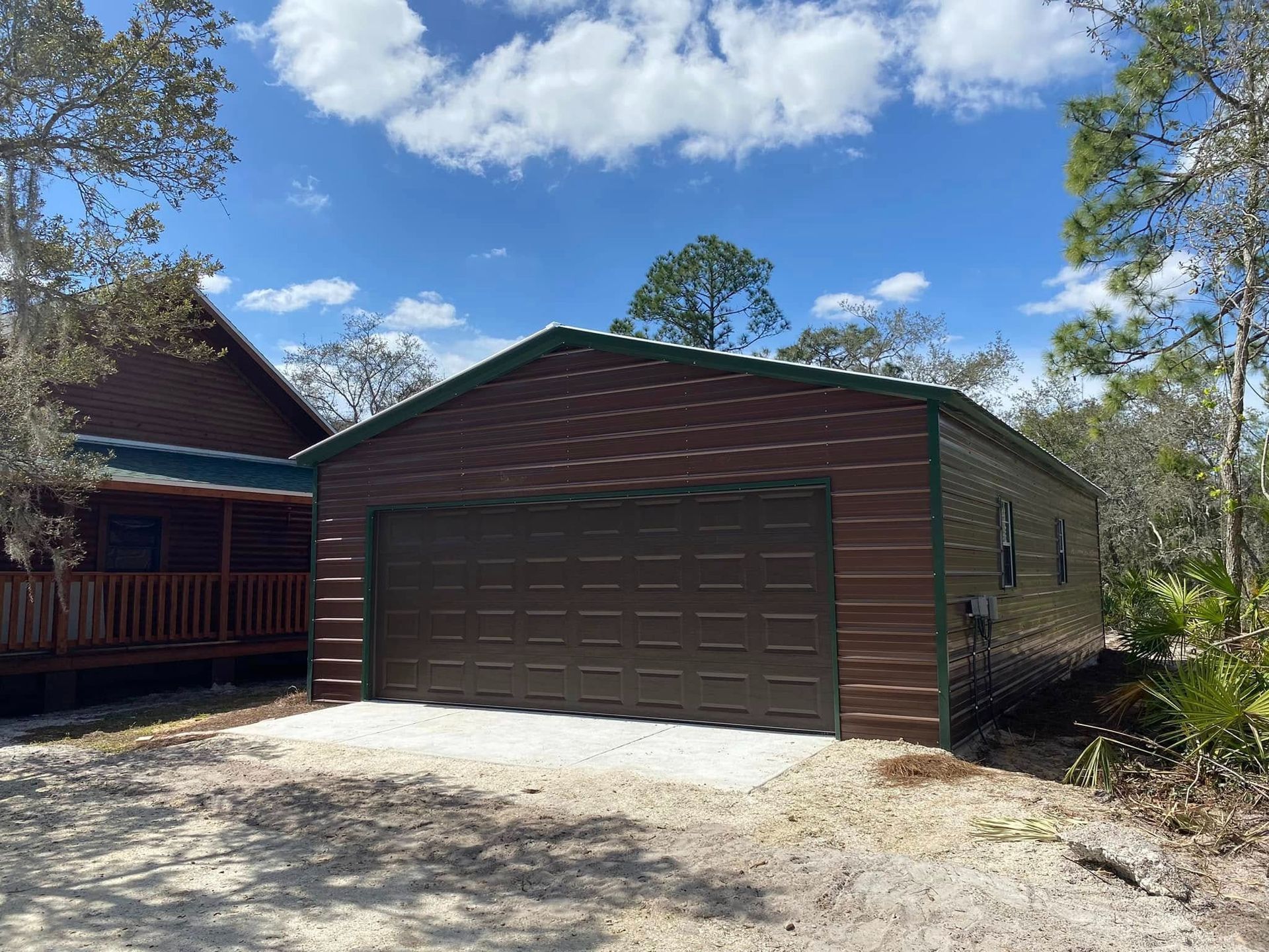 A brown metal garage with a dark door sits next to a wooden cabin in a wooded, sunny setting.