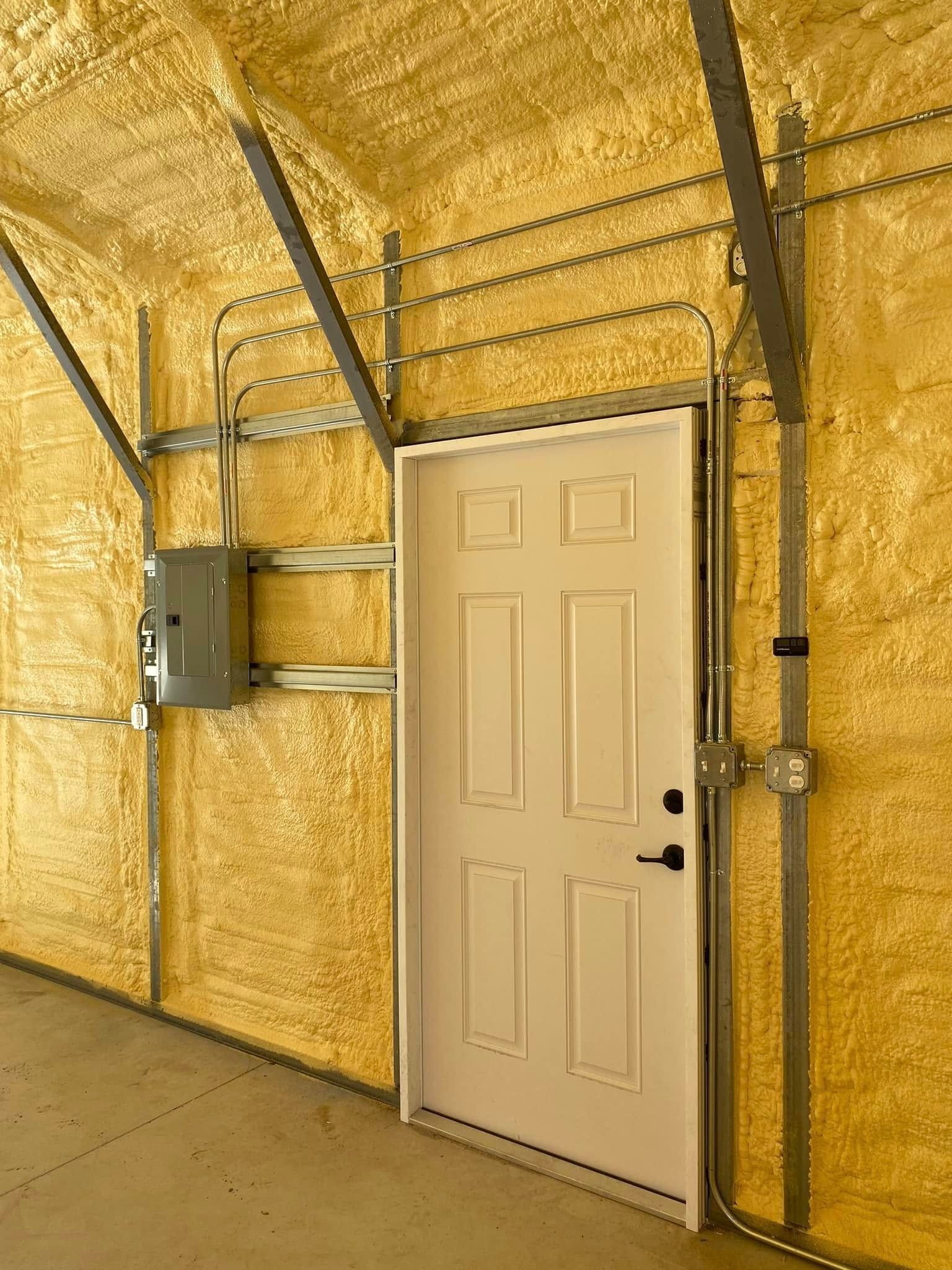 Interior of a metal building featuring a white door, an electrical panel, and conduit pipes against yellow foam insulation.