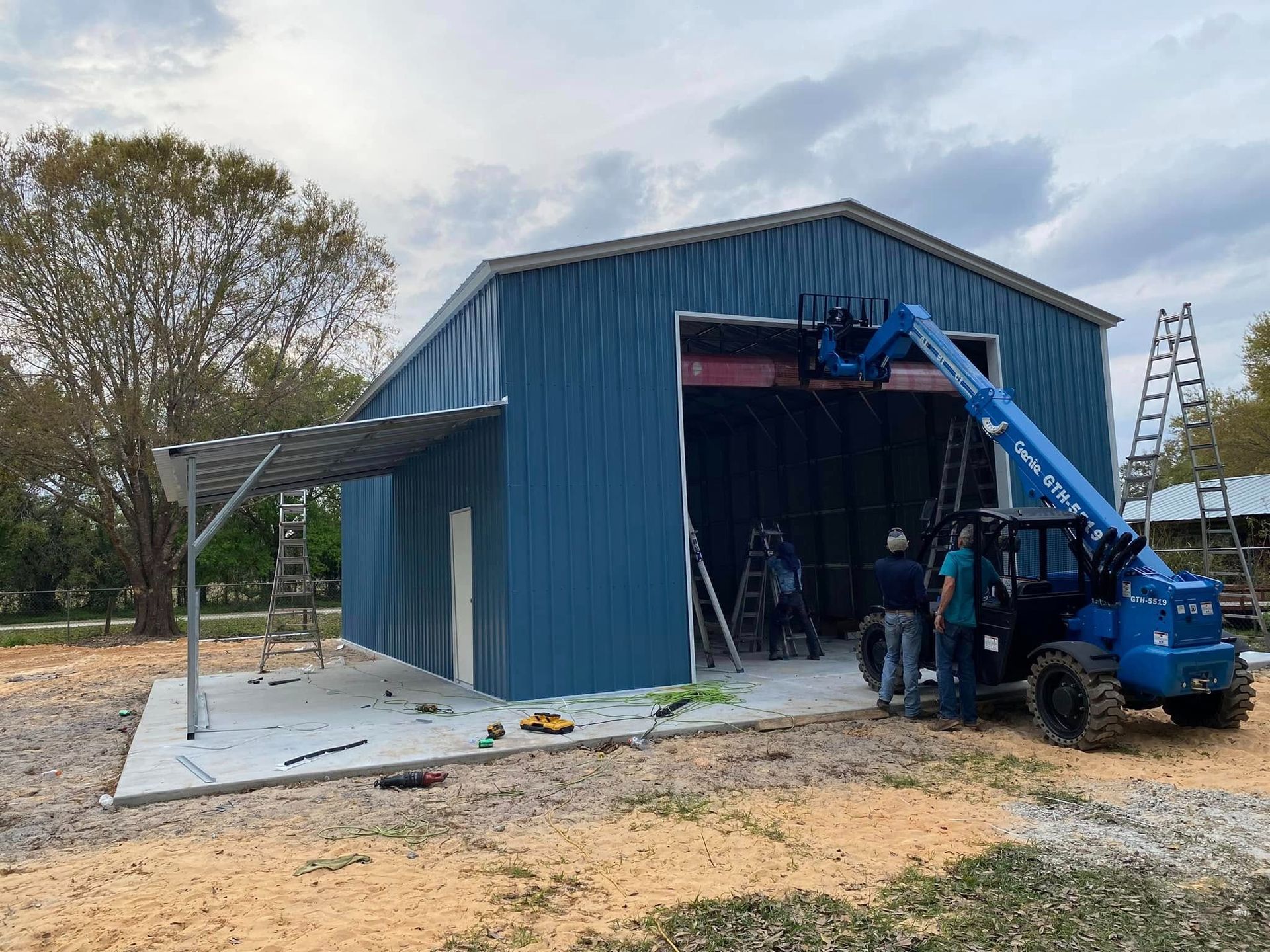 Construction crew using a blue telehandler to work on the entrance of a blue metal building with an attached carport.