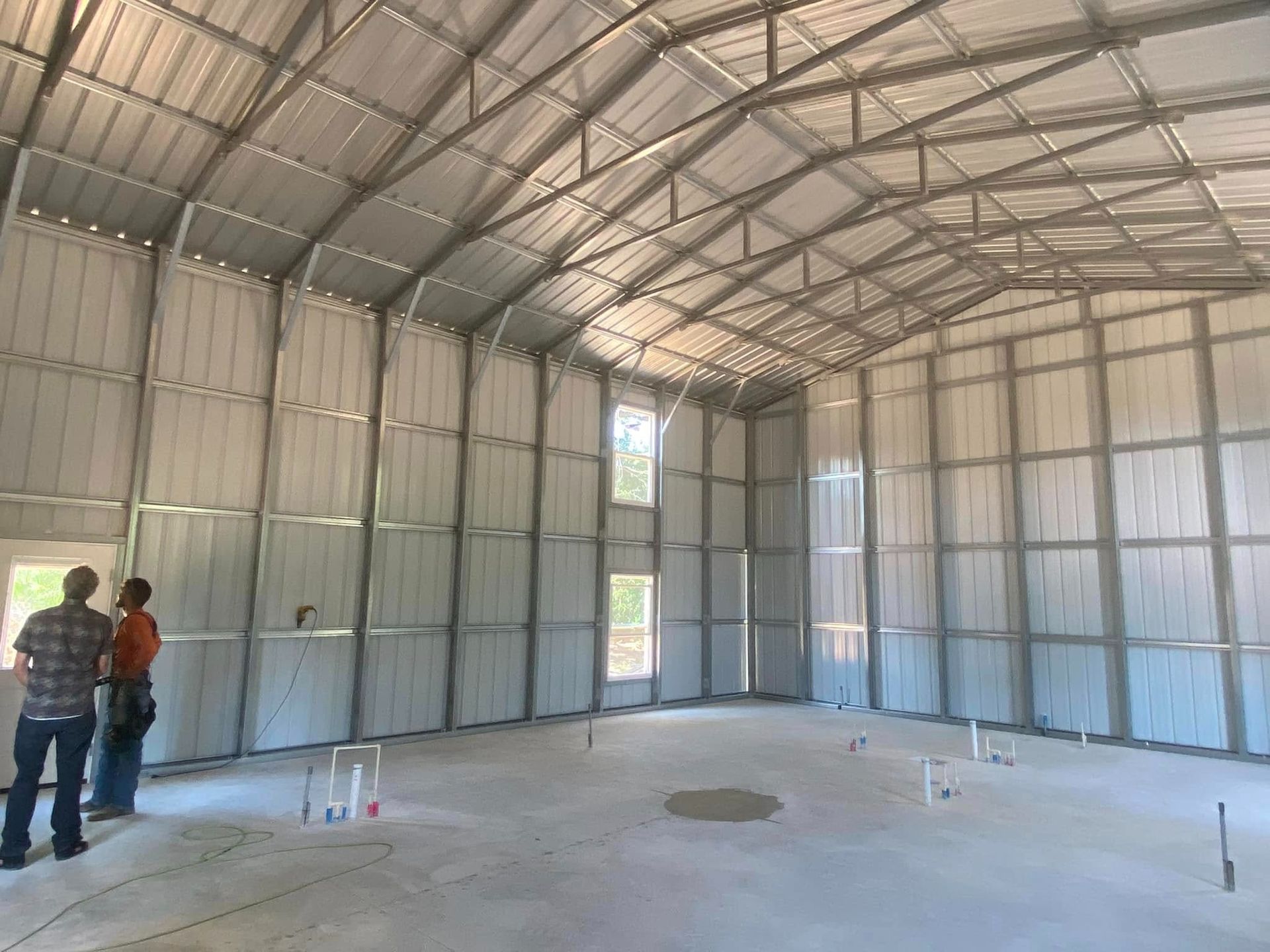 Two people stand inside a large, unfinished metal building with insulated walls and an open, steel-trussed ceiling.