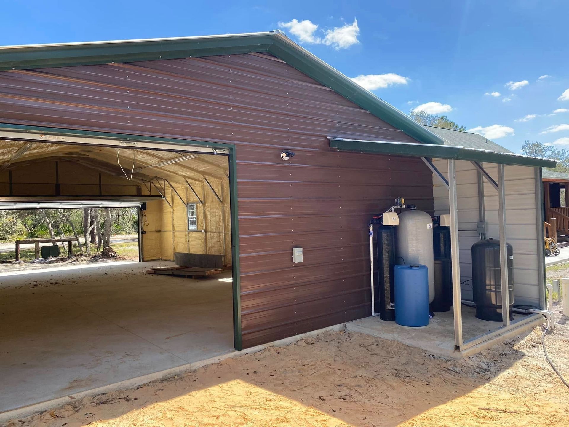 A metal garage with an open bay next to a covered area housing water filtration tanks on a sandy, sunny lot.