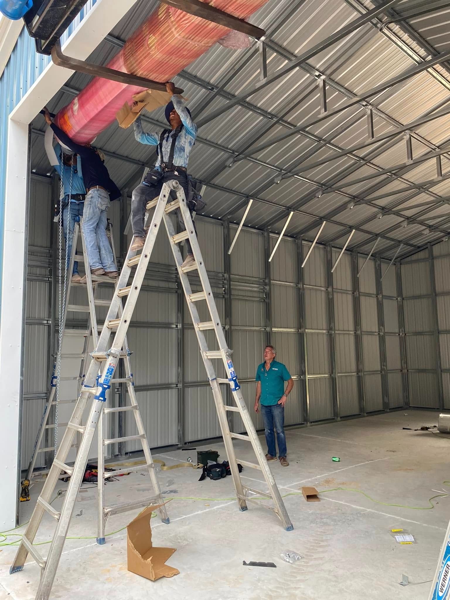 Workers install a large roll-up door in a metal warehouse, using ladders to reach the upper framework.