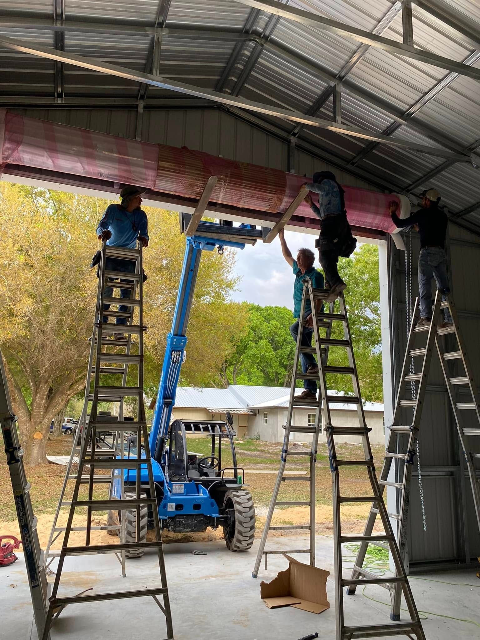 Three people on ladders work to install a large horizontal beam above an open doorway in a metal building.