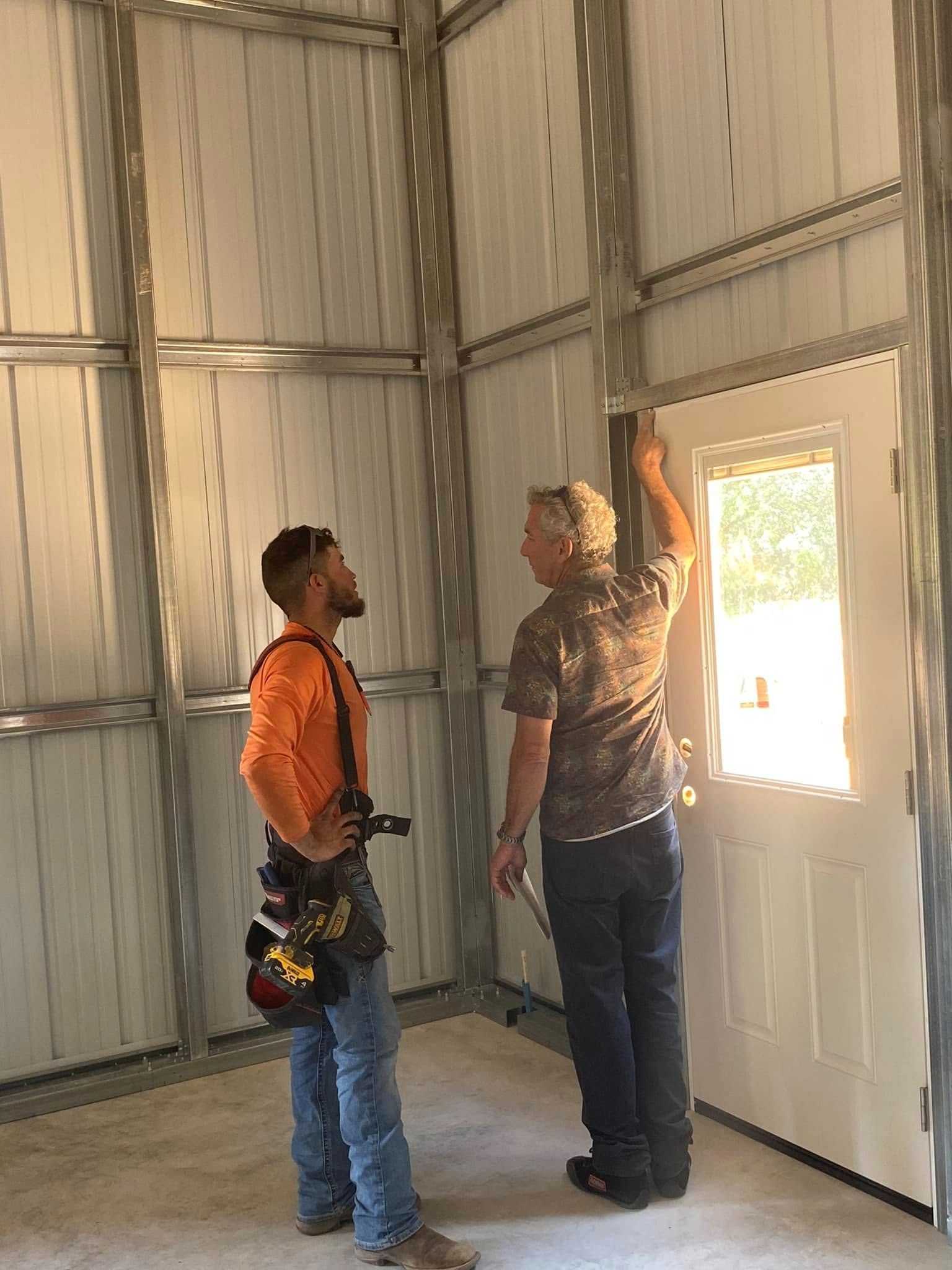 Two workers in a metal-framed structure talk while standing near an installed white door.