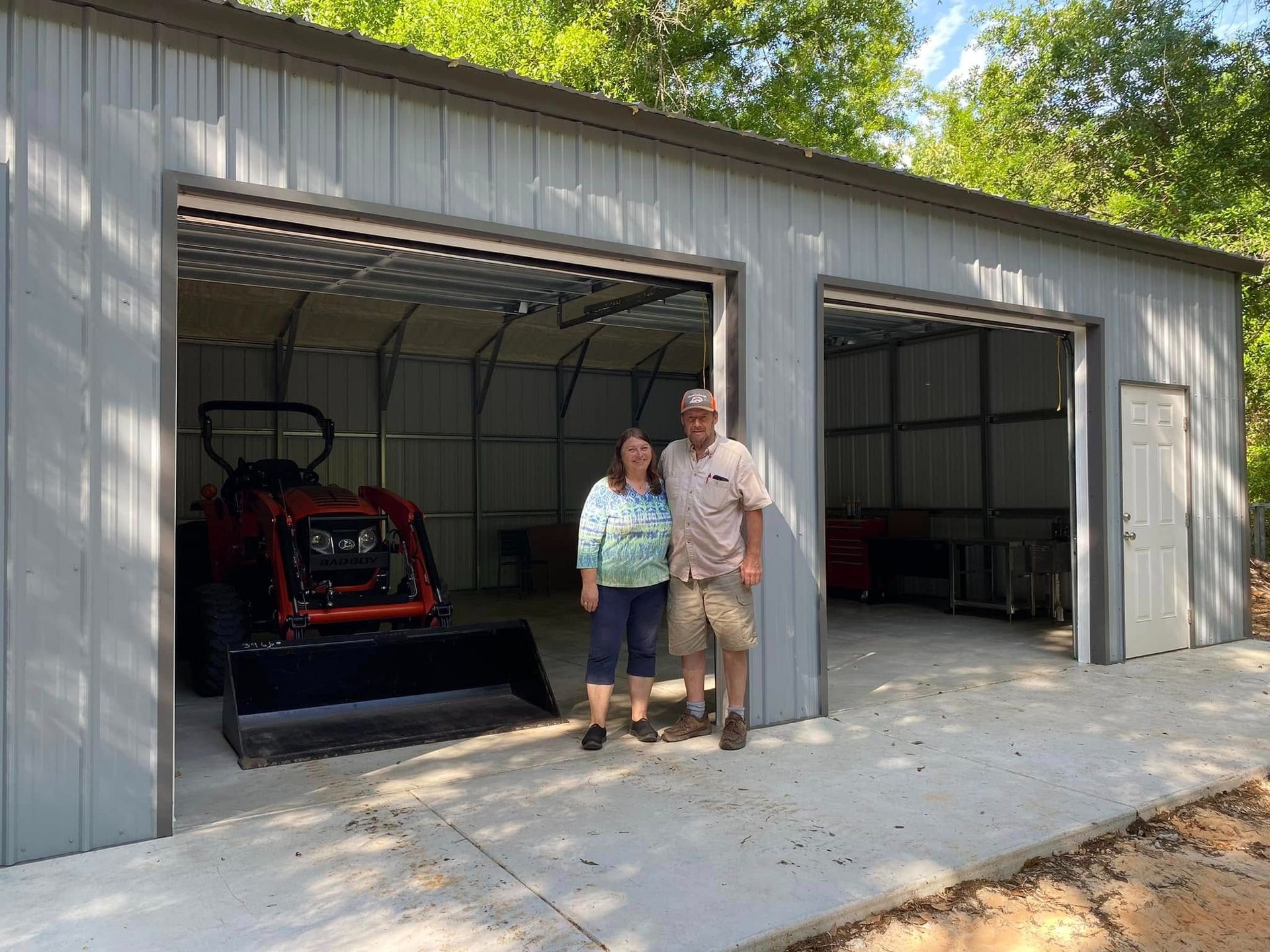 A smiling couple stands in front of a new gray metal garage, with a red tractor parked in the open left bay.