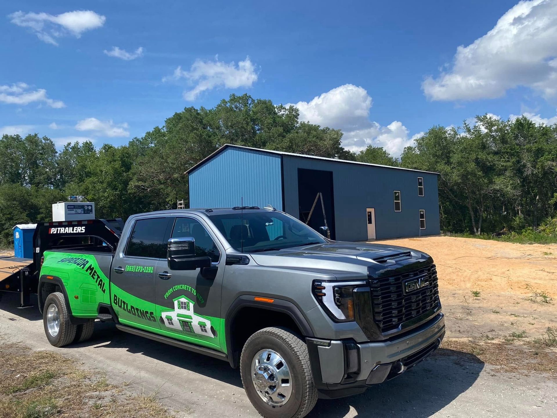 A gray GMC dually pickup truck with bright green decals parked on a gravel path in front of a blue metal industrial shed.