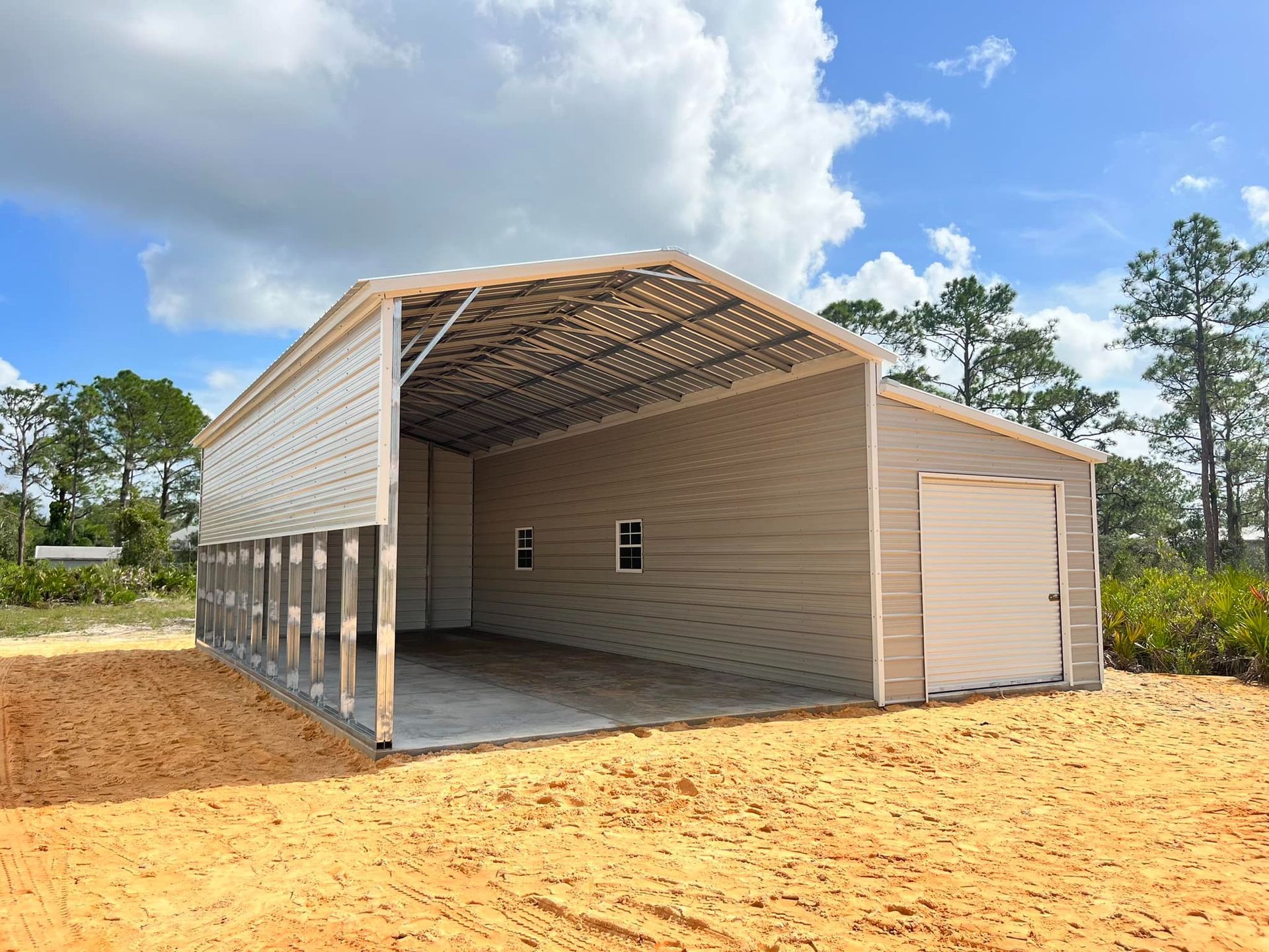 A tan metal garage with an open bay, a closed roll-up door, and two small windows on a sandy, rural lot.