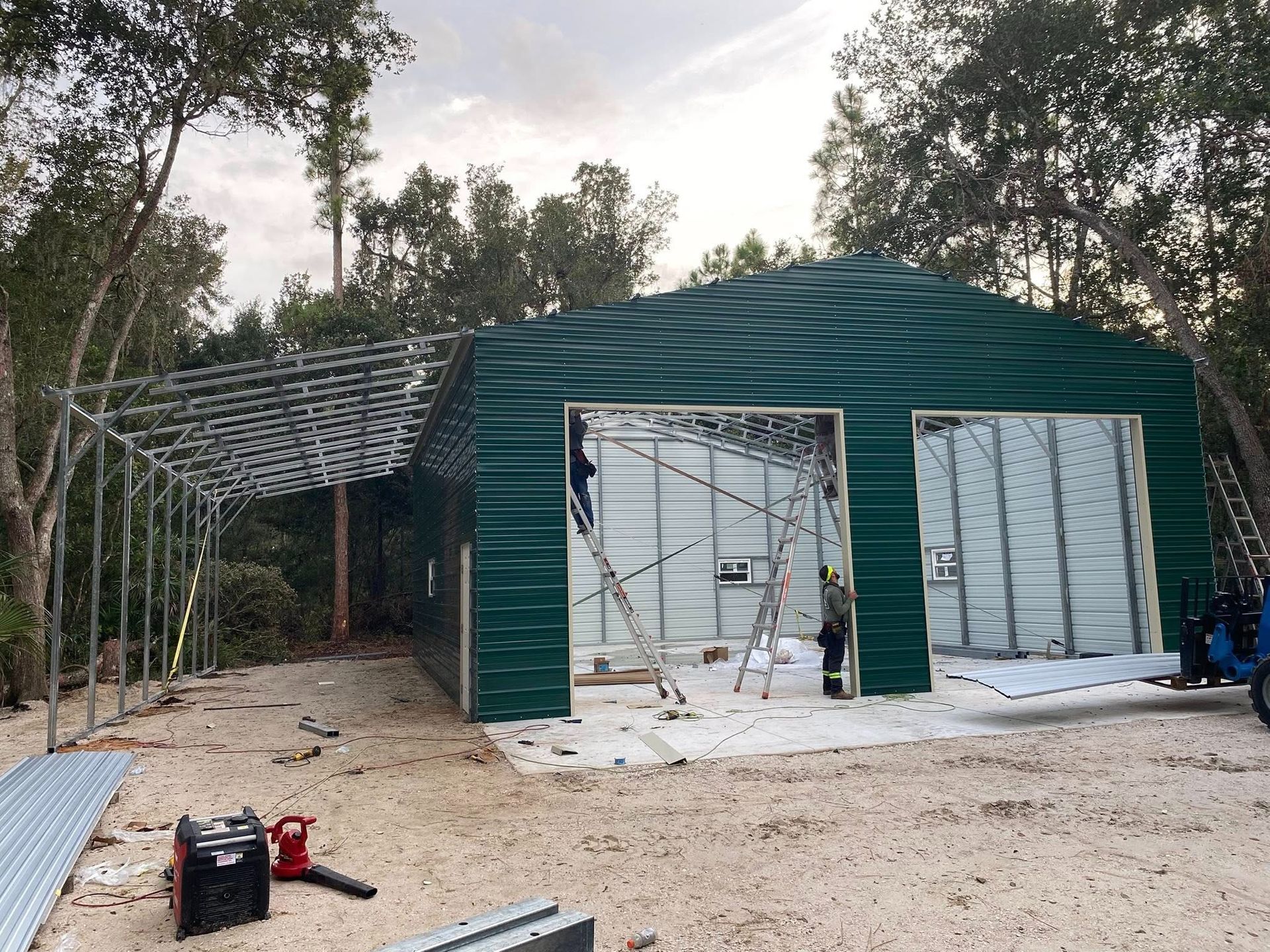 Workers install green metal siding on a large warehouse frame with an attached metal carport in a wooded lot.