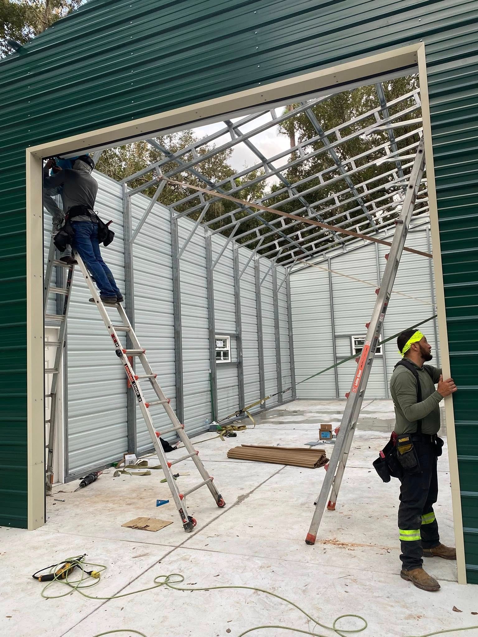 Two construction workers install metal siding on a building frame; one climbs a ladder, the other stands on the ground.