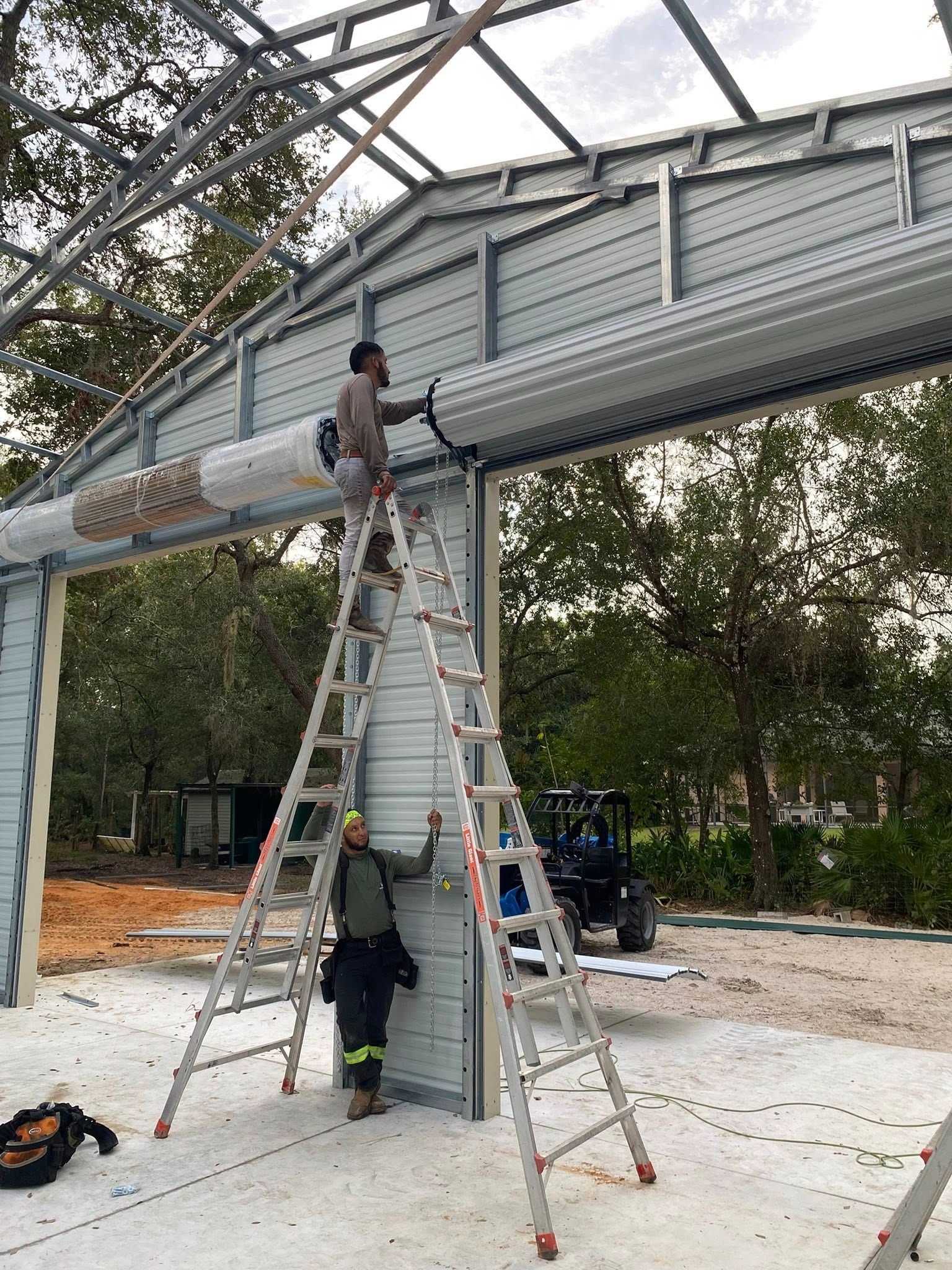 Two workers install a large white rolling door mechanism on a steel-framed building under construction.