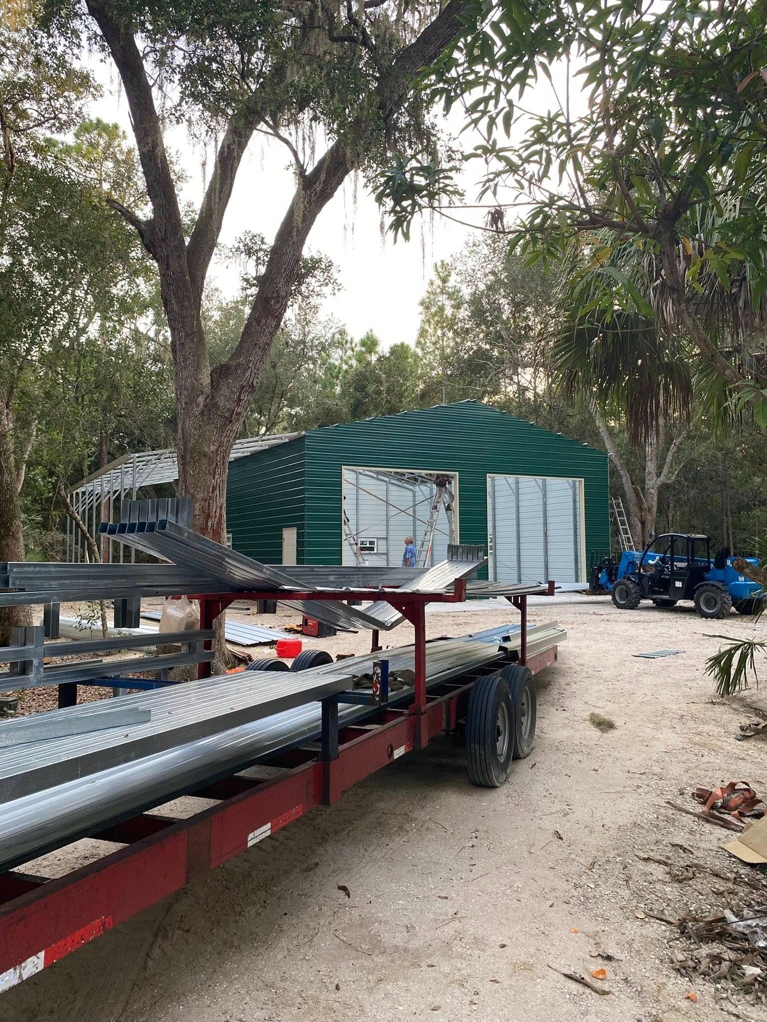 A red flatbed trailer loaded with steel beams parked in front of a green metal workshop surrounded by trees.