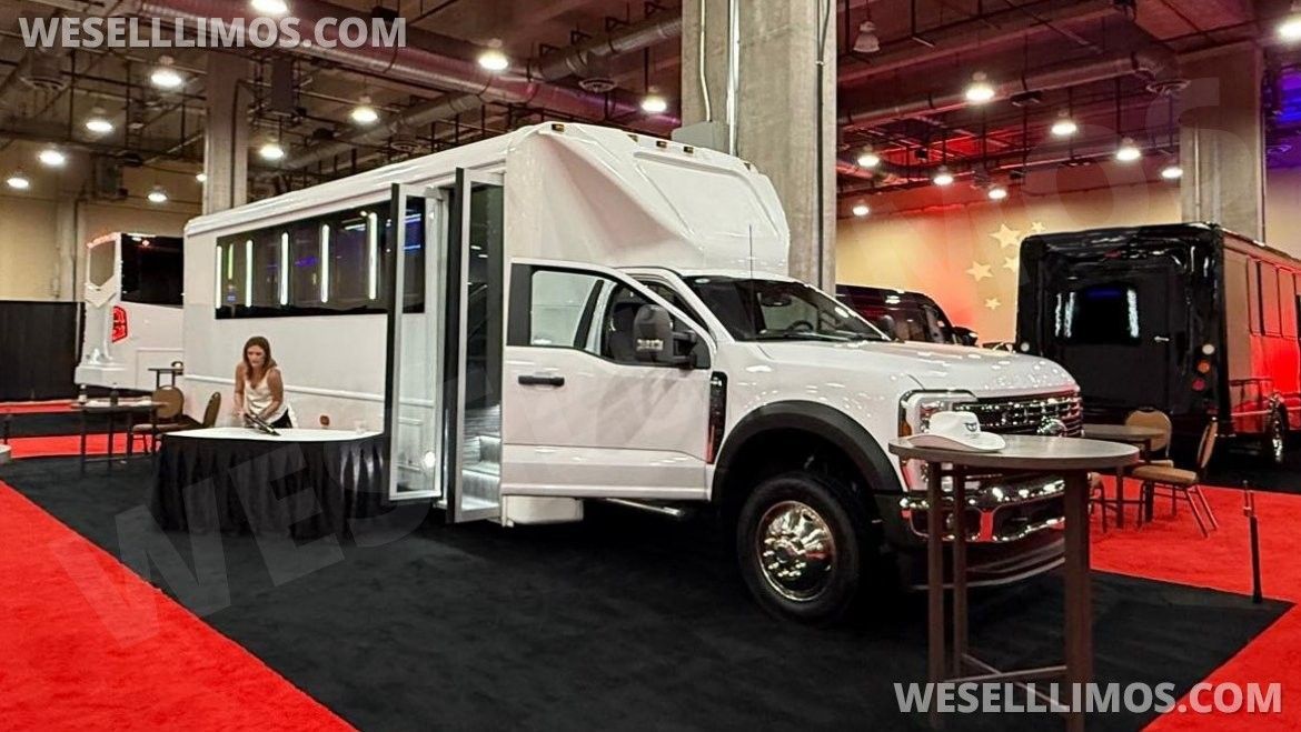 White limo bus with open door parked at a show, a woman sits at a table near the entrance.