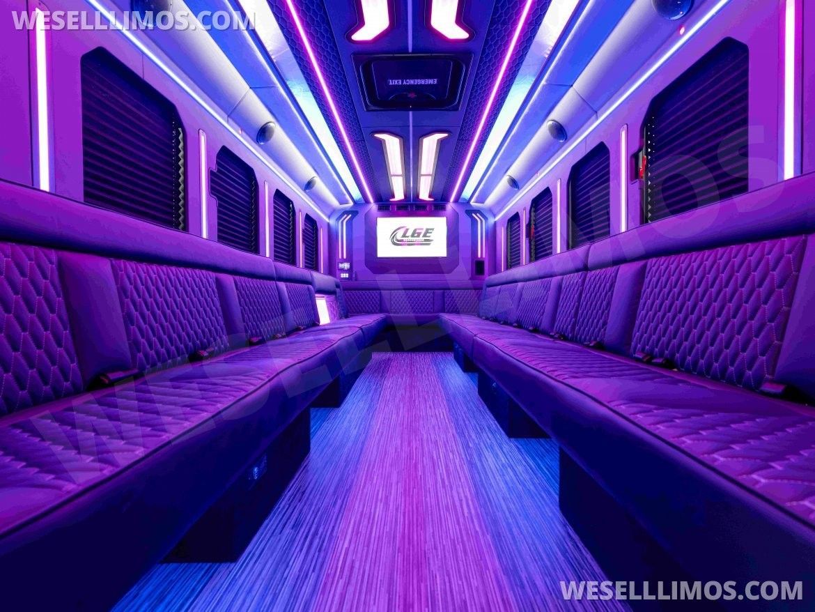 Interior of a party bus with purple lighting, two rows of cushioned seating, and a central aisle.