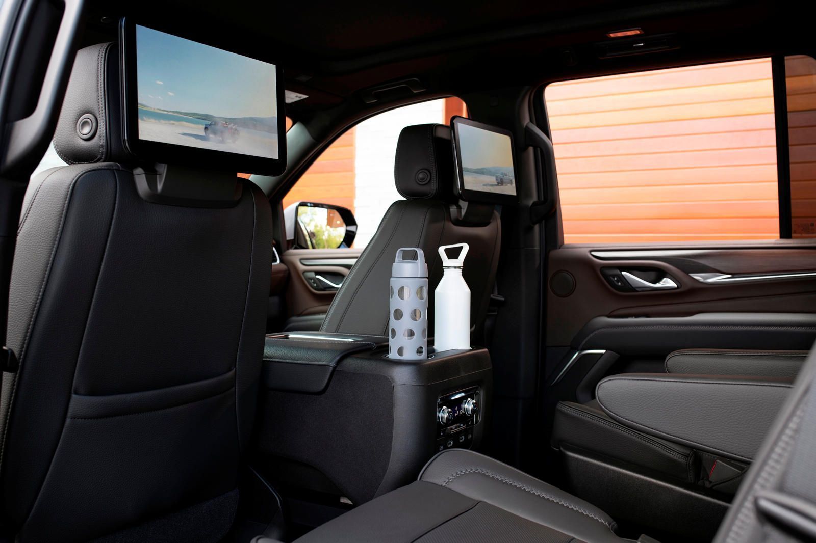 Rear interior of a vehicle with two screens, drinks, and black seats.