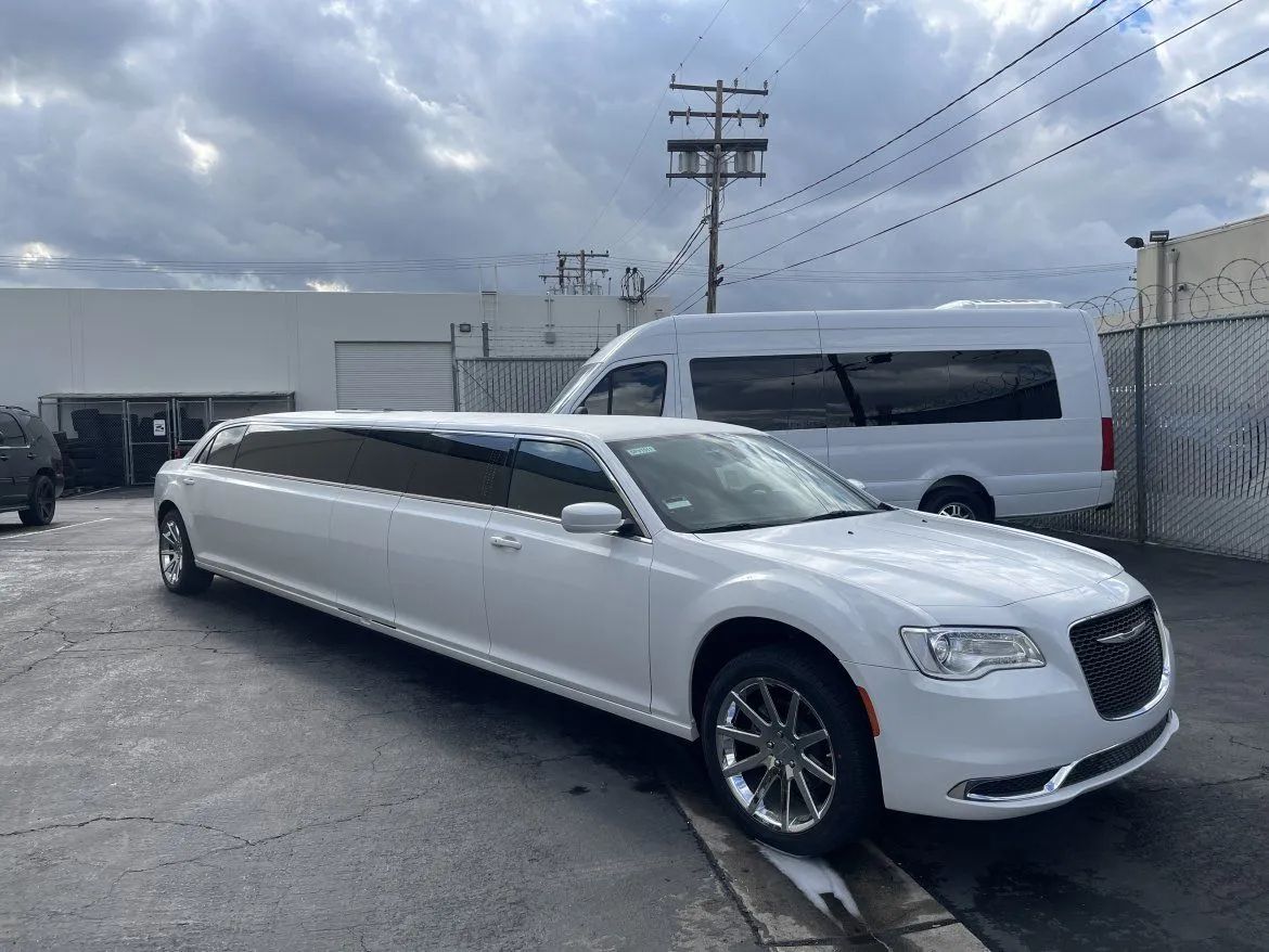 White stretch limousine parked in an industrial lot beside a white van and chain-link fence