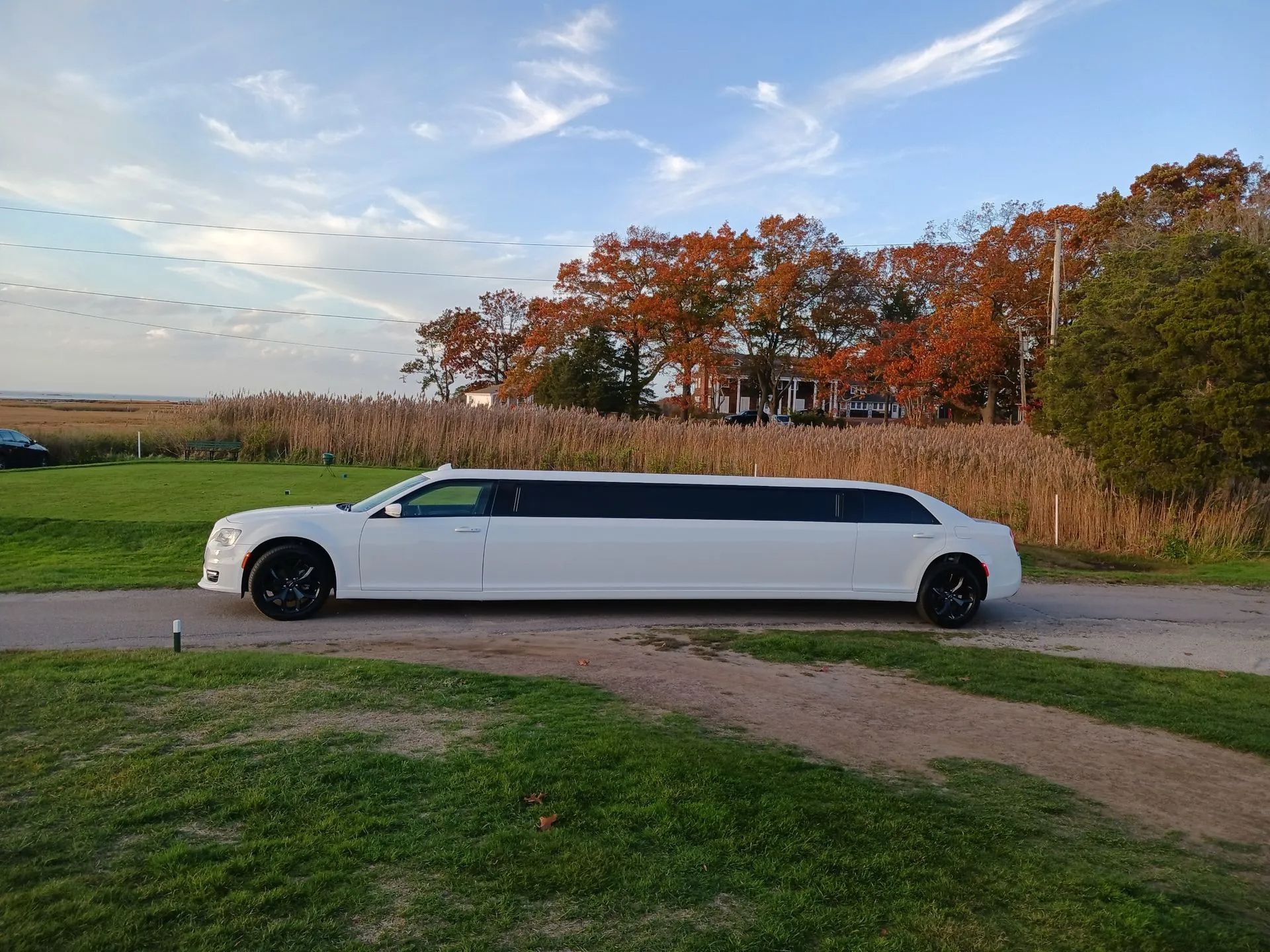 White stretch limousine parked on a country road beside green fields and autumn trees under a blue sky