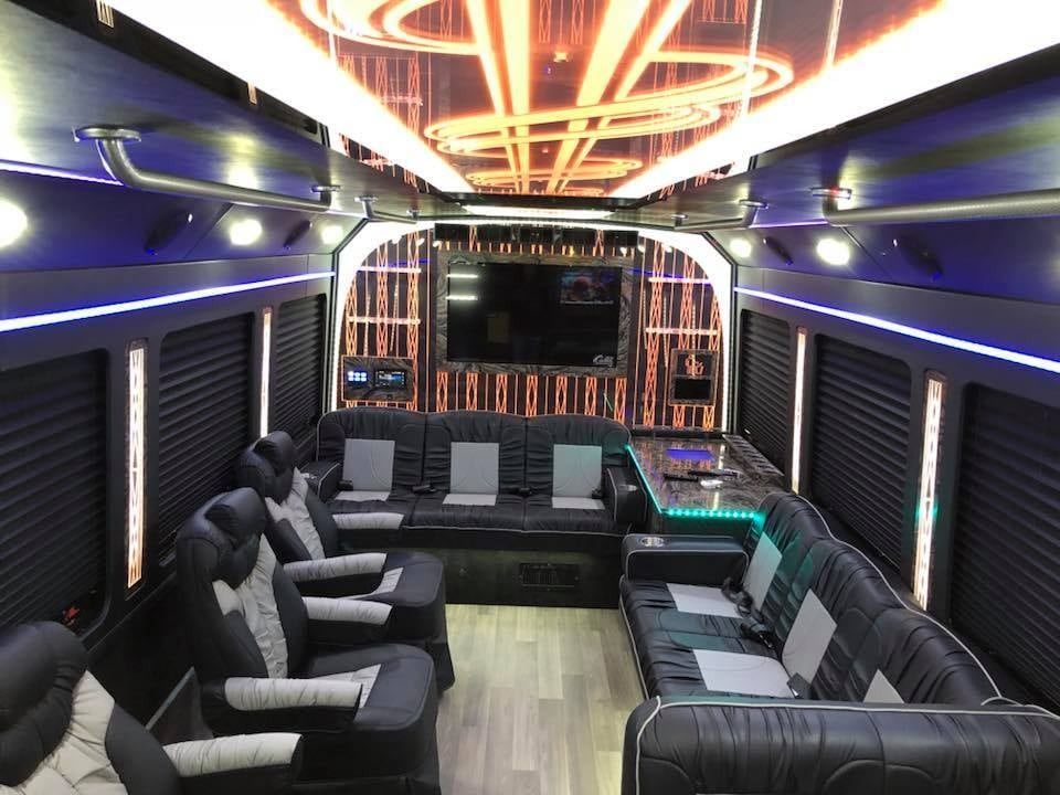 Interior of a luxury party bus with black and gray seating, a large TV, and colorful lighting.