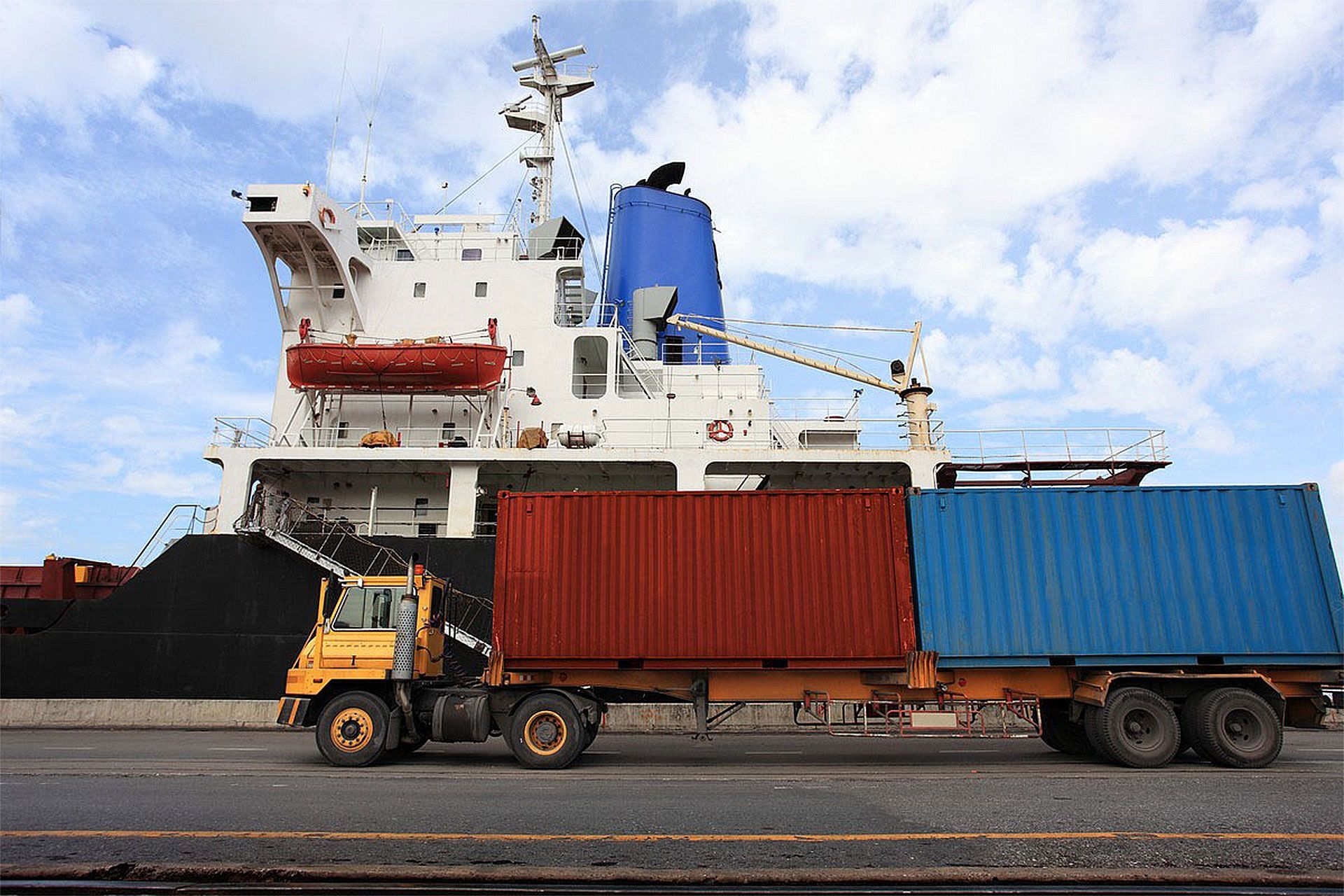 Forklift truck carrying two shipping containers at a dock, with a cargo ship in the background.