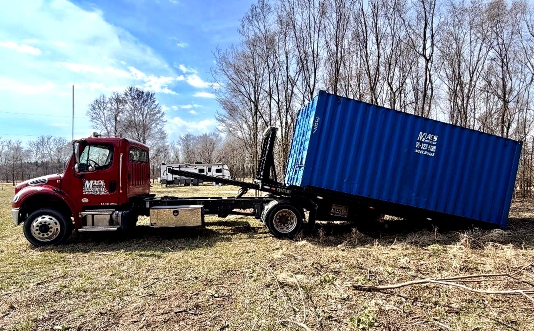 Forklift truck carrying two shipping containers at a dock, with a cargo ship in the background.