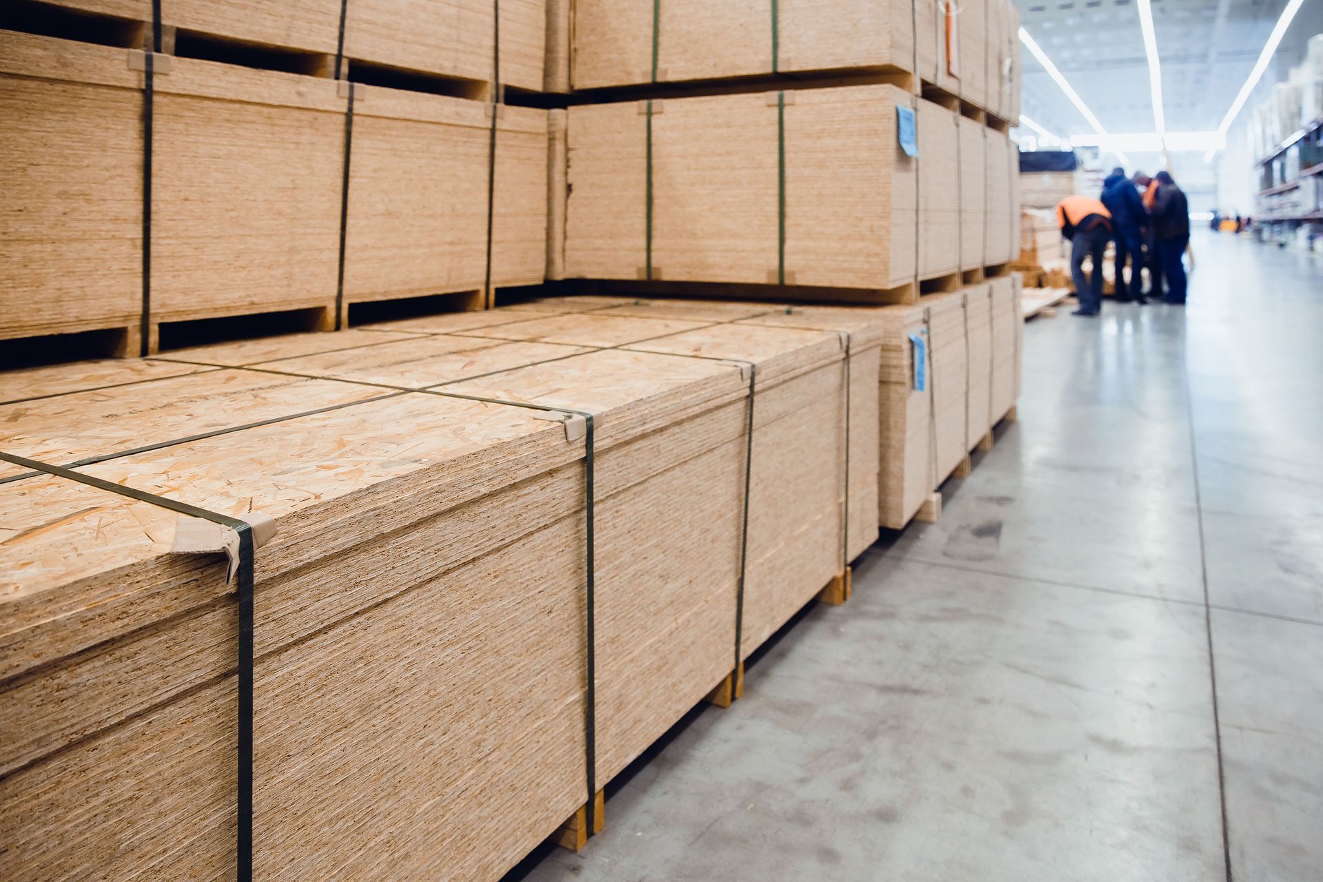 Piles of stacked particleboard secured with straps in a warehouse; several people in the background.