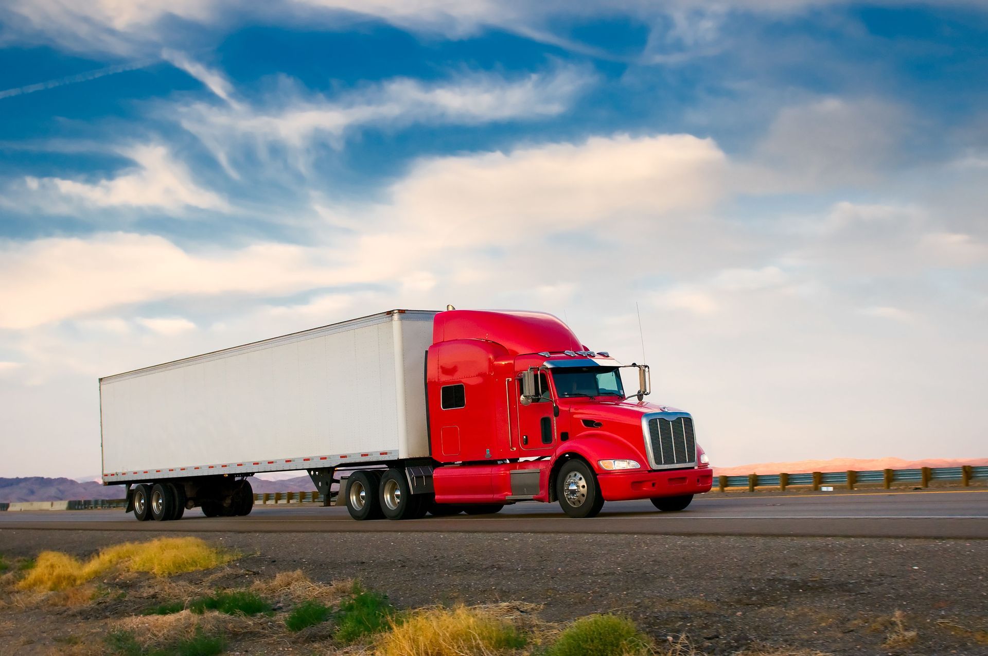 Red semi-truck with white trailer driving on a highway under a cloudy blue sky.